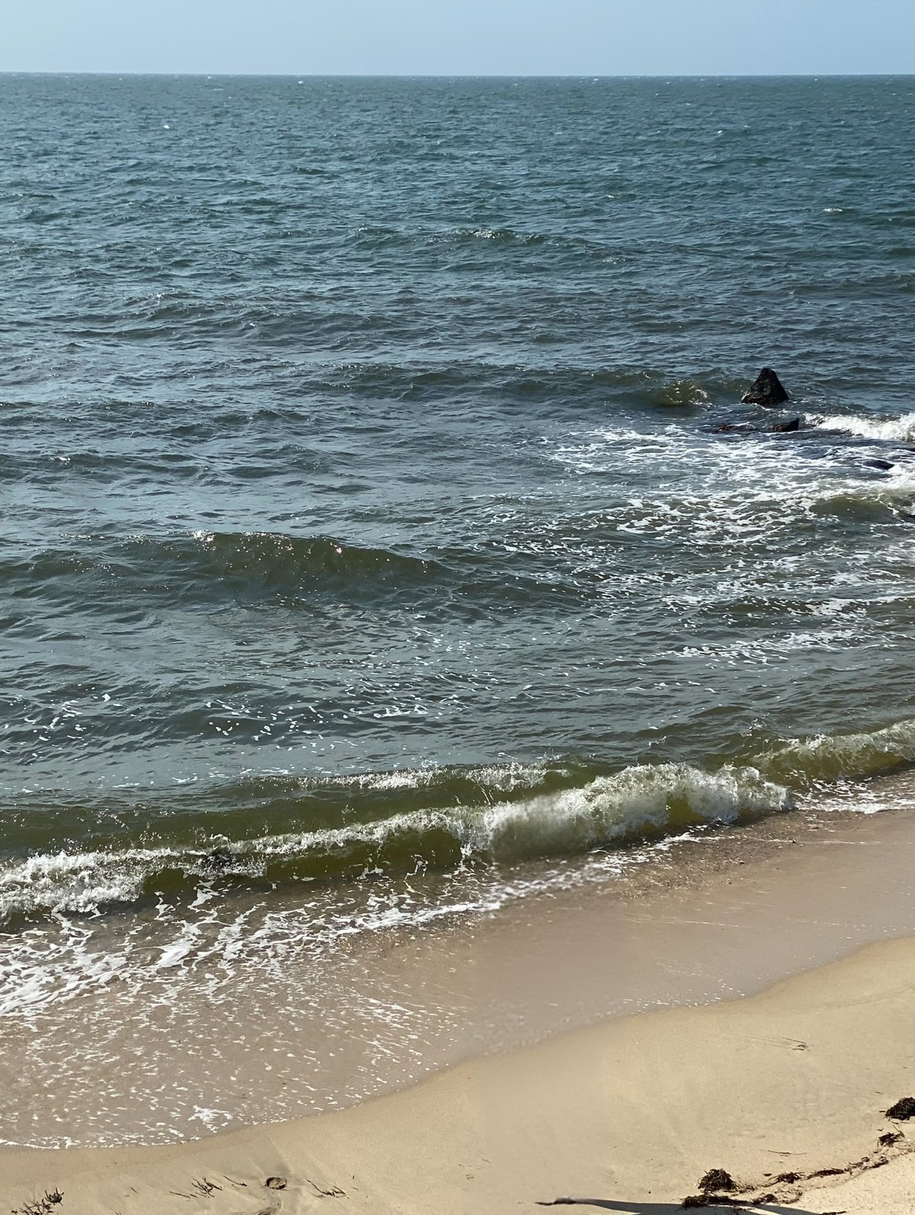 Ocean waves crashing onto sandy beach with rocks in the water and blue sky above.