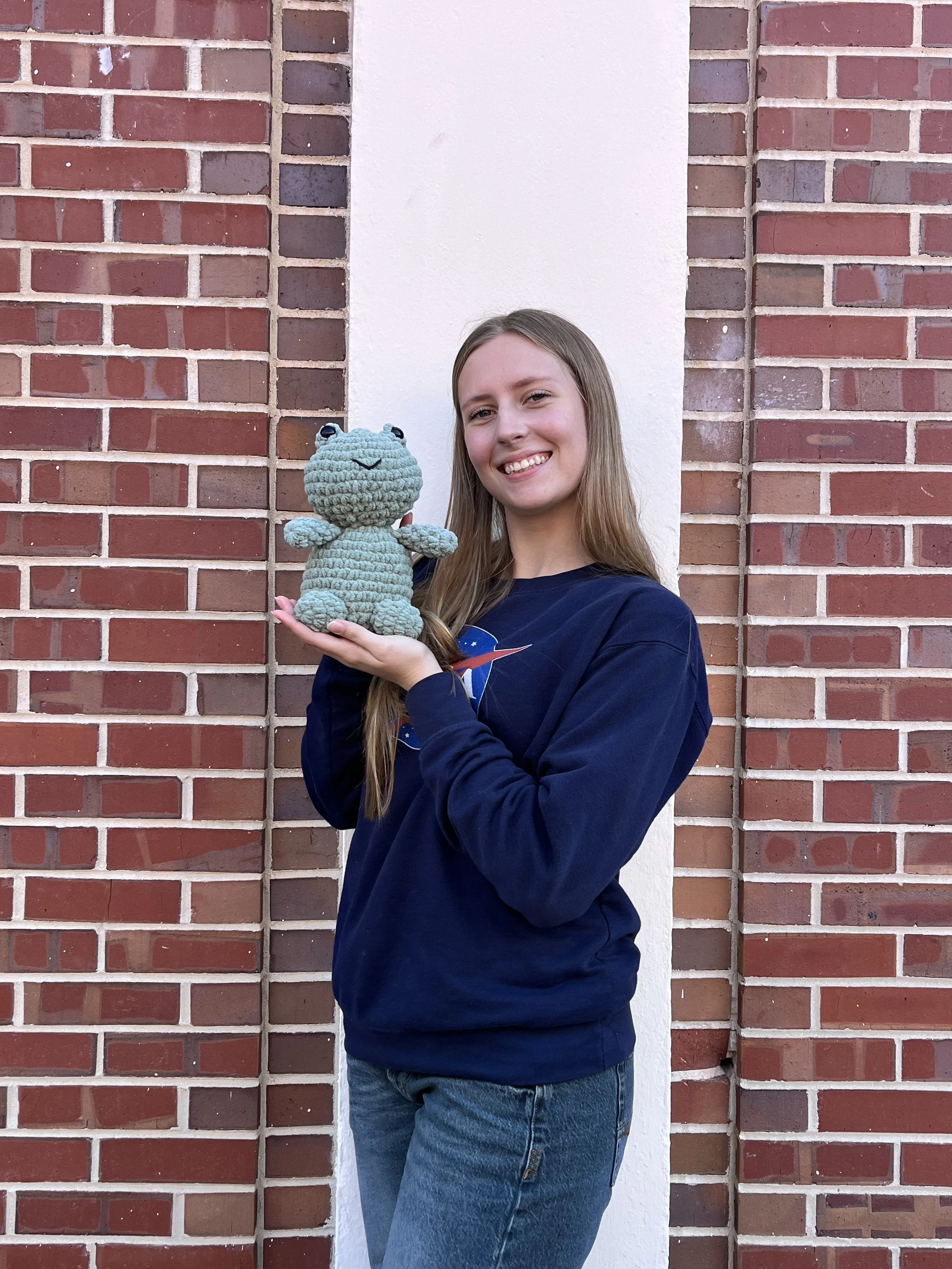 A young woman with long blonde hair and a navy blue sweatshirt holding a crocheted stuffed frog and smiling at the camera.