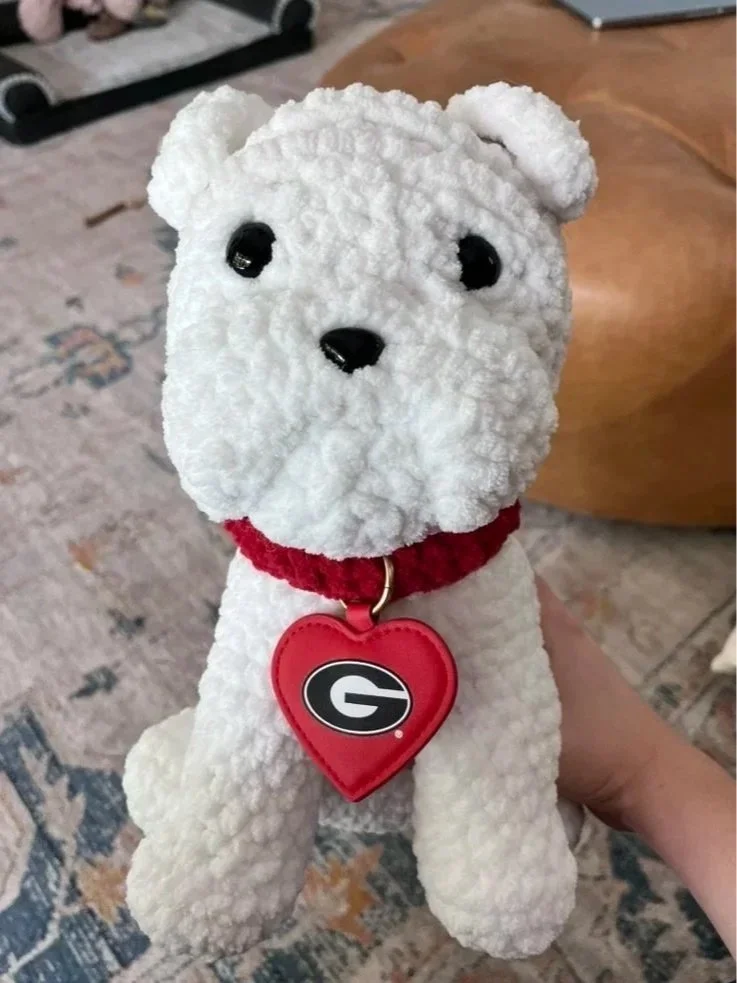 A plush toy stuffed animal resembling a dog, with a white textured body, black eyes, and a black nose, wearing a red collar with a heart-shaped tag with a 'G' logo.