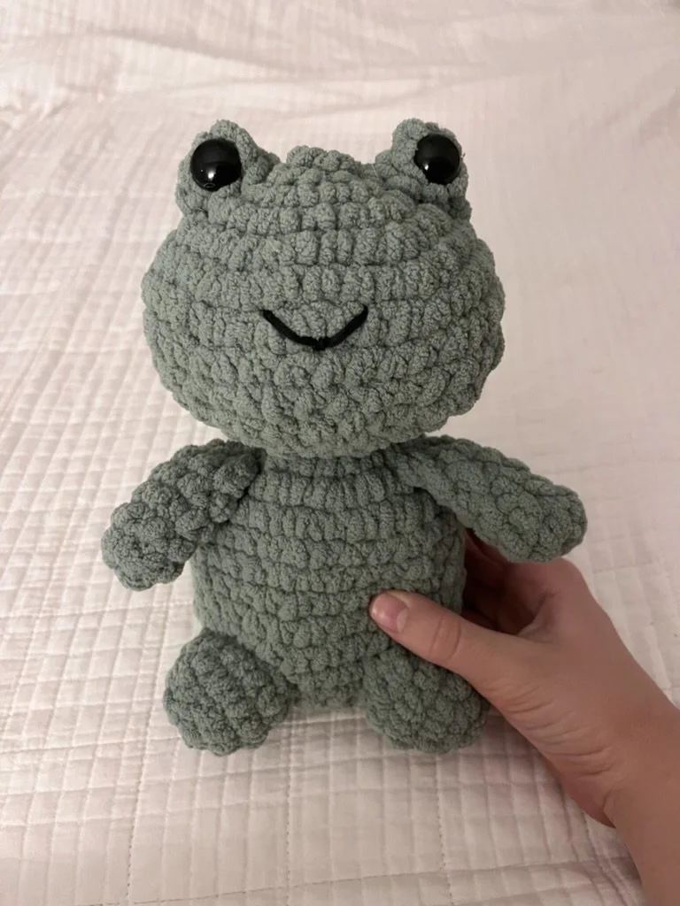 A plush toy resembling a frog with textured green fabric, large black eyes, and a small smiling mouth, being held by a person in front of a pink quilted background.