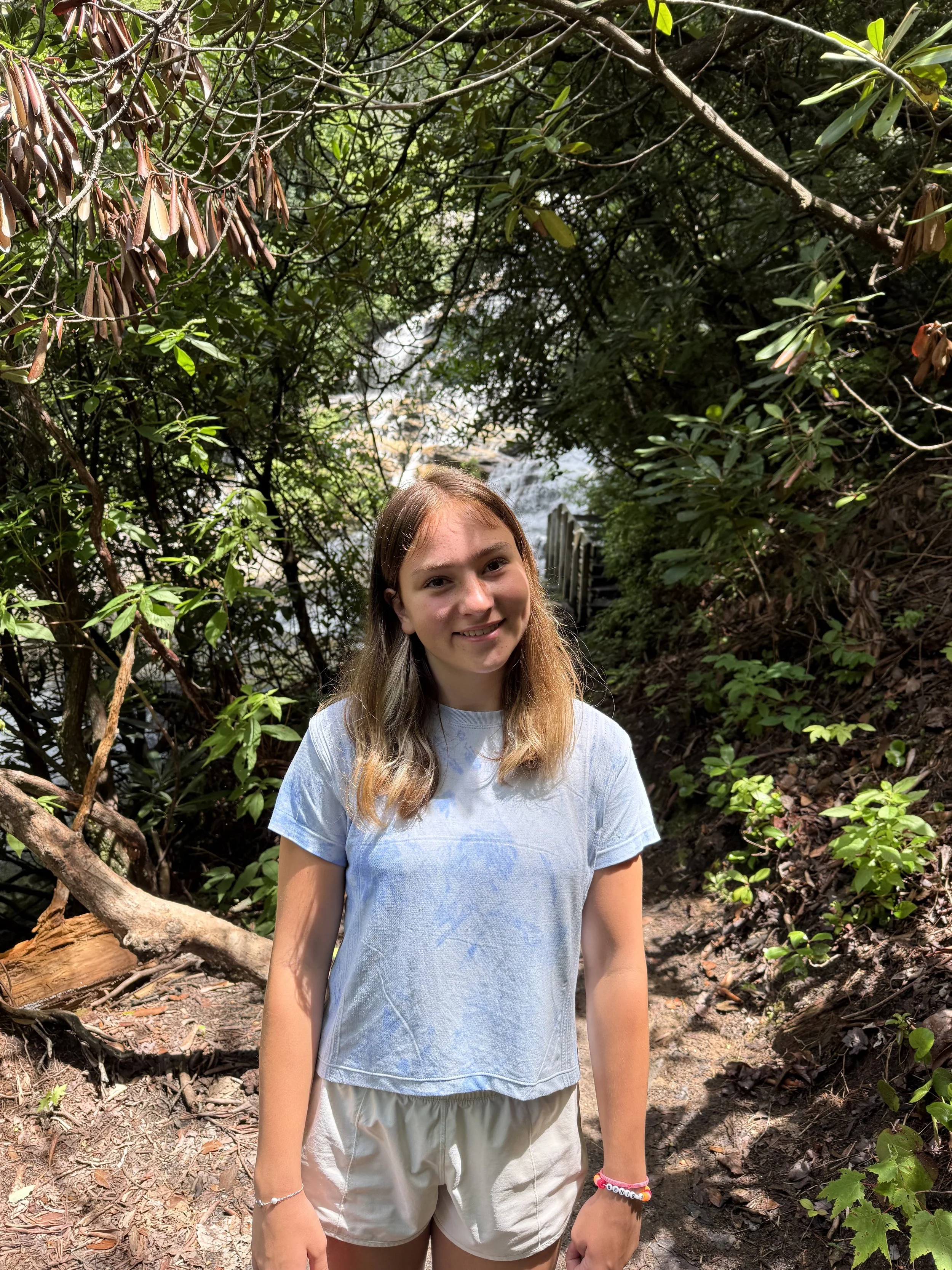 A young woman standing in a lush, green forest near a stream, smiling at the camera.