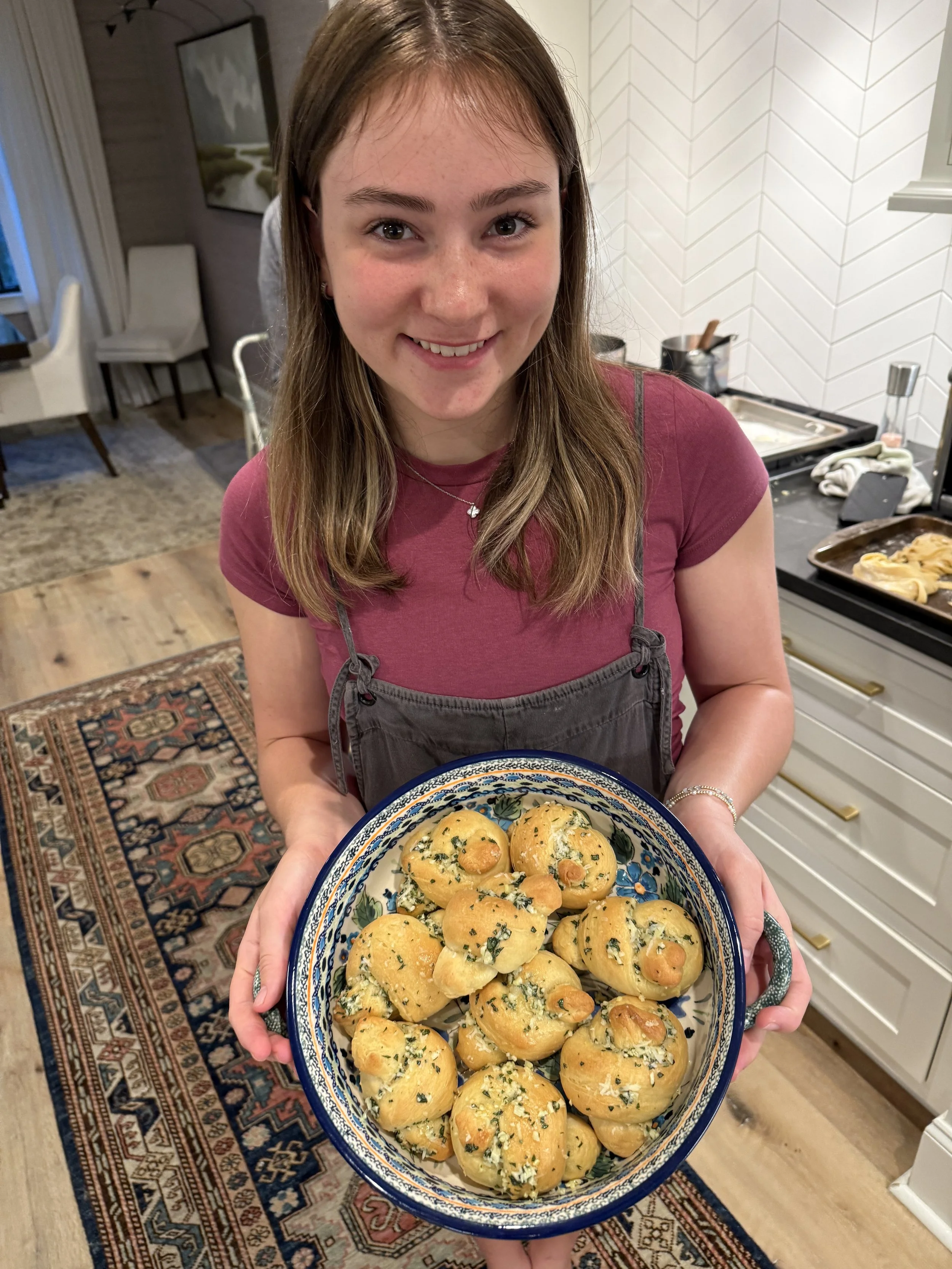 A young woman with light brown hair, wearing a maroon shirt and gray apron, smiling while holding a large plate of garlic knots garnished with chopped herbs in a kitchen.