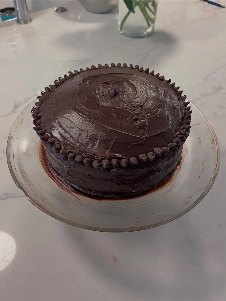 Chocolate cake with frosting and decorative chocolate dots around the top edge, placed on a glass cake stand.