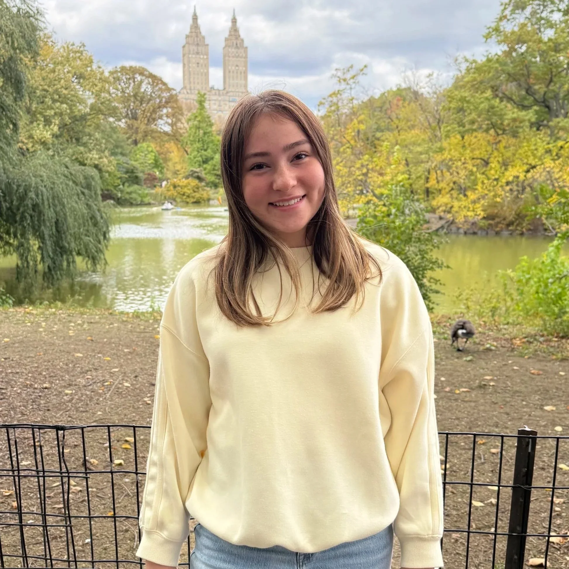 A young woman smiling outdoors near a pond with autumn trees, and a city skyline with tall buildings in the background, wearing a cream-colored sweatshirt.