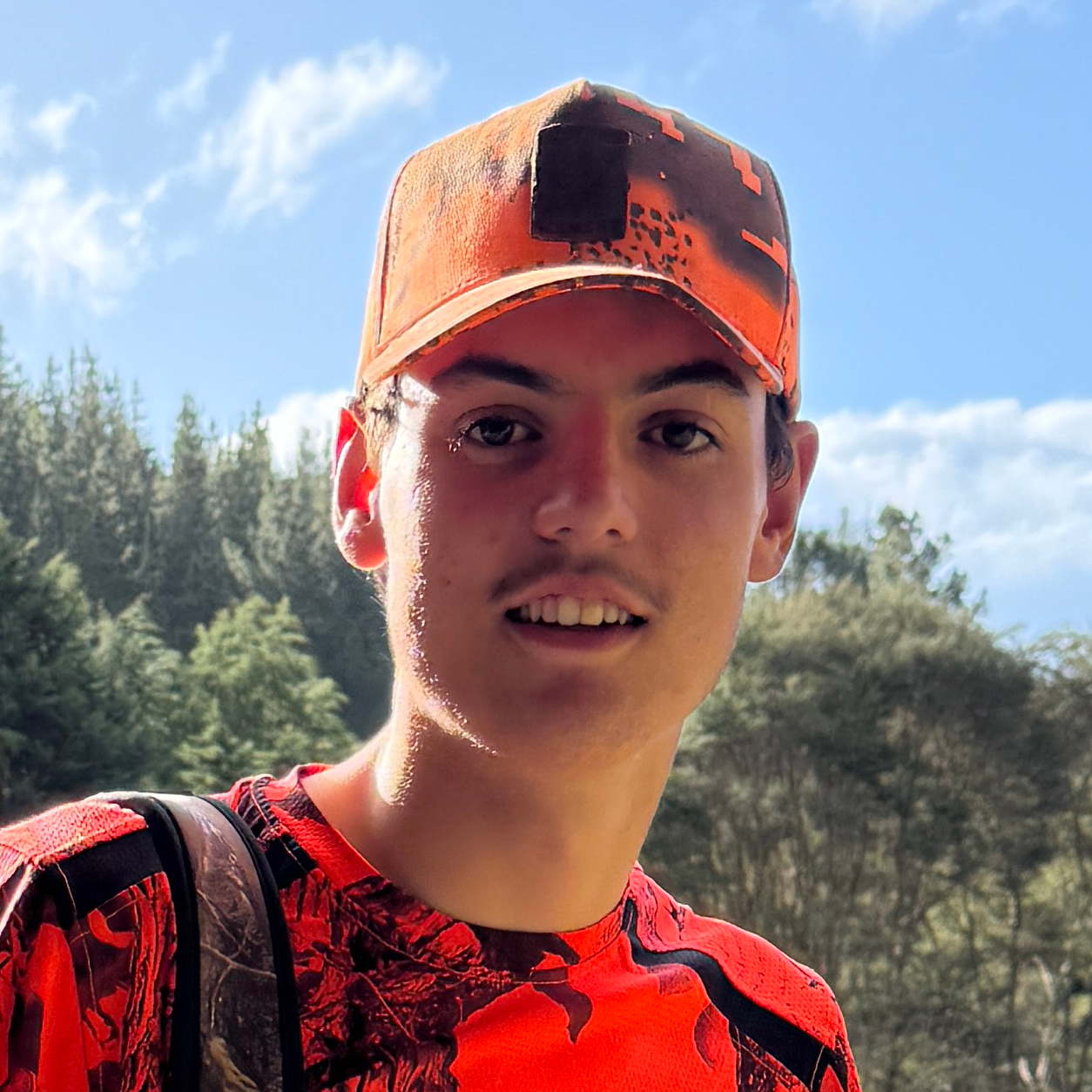 Young man smiling outdoors in a forest, wearing an orange camouflage shirt and a matching cap.