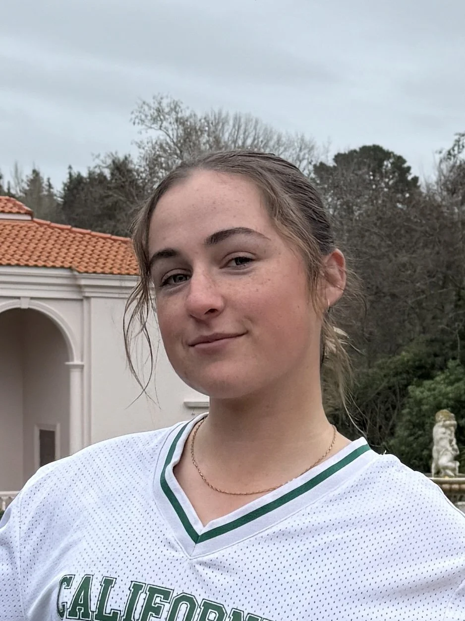 A man with curly dark hair, glasses, and facial hair looking to the side while sitting indoors.