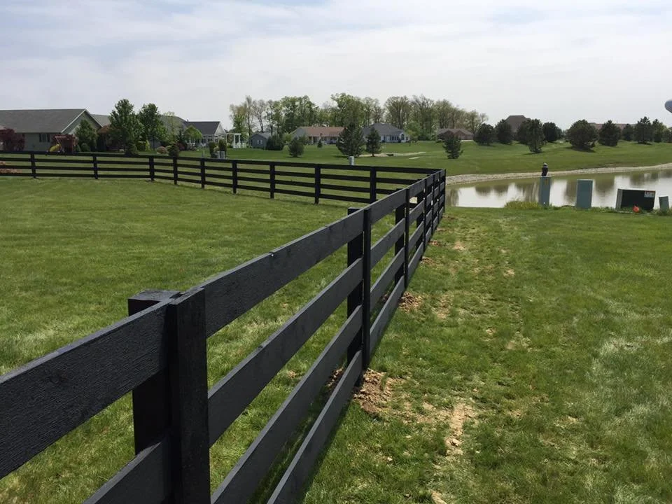 Black wooden fence running along the grassy edge of a pond in a suburban neighborhood with houses and trees in the background.