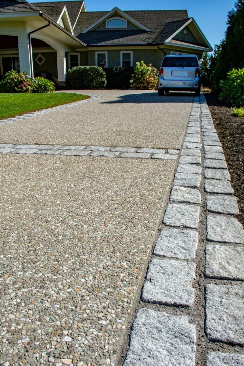 Residential house with a gravel driveway, bordered by stone pavers, parked car in driveway, landscaped garden with bushes and flowers, clear blue sky.
