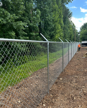 Construction site with a long chain-link fence and a person in an orange shirt in the background, trees on one side, and a dirt path on the other.
