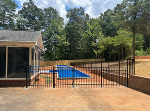 A backyard with a swimming pool surrounded by a black metal fence and a paved area, with trees and a house in the background.