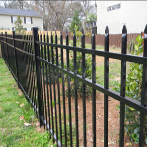 Black metal fence with wooden tops in a yard with some plants and grass, adjacent to a white building and neighboring houses.