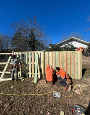 Workers building a wooden fence outdoors with tools and safety gear on a clear day.