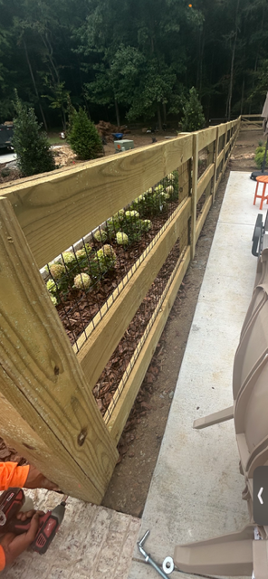 Wooden fence with wire garden trellis, planted with leafy green plants, next to a concrete sidewalk and outdoor seating area, with trees and parked cars in the background.