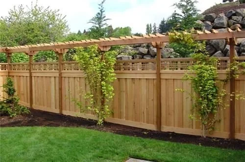 A wooden privacy fence with vertical slats and a lattice top, surrounded by green trees and bushes, with a grassy lawn in front.