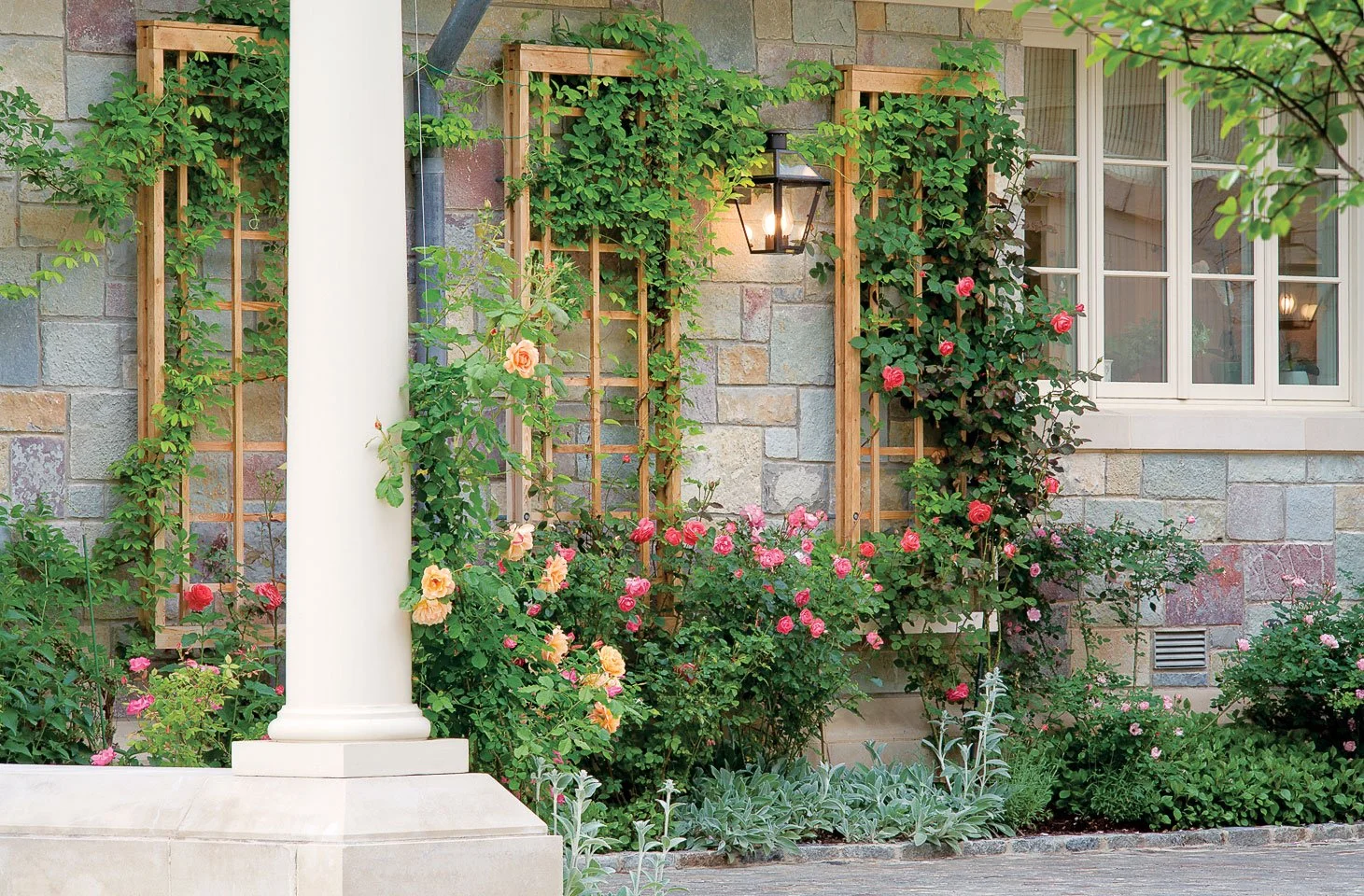 Flowering garden near a house with a brick exterior, wooden trellises, and a window with white trim, illuminated by a wall-mounted lantern.