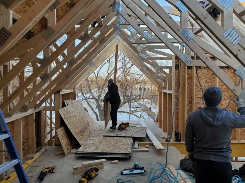 Two workers are constructing the interior of a house, installing roof trusses, with construction materials and tools scattered around.