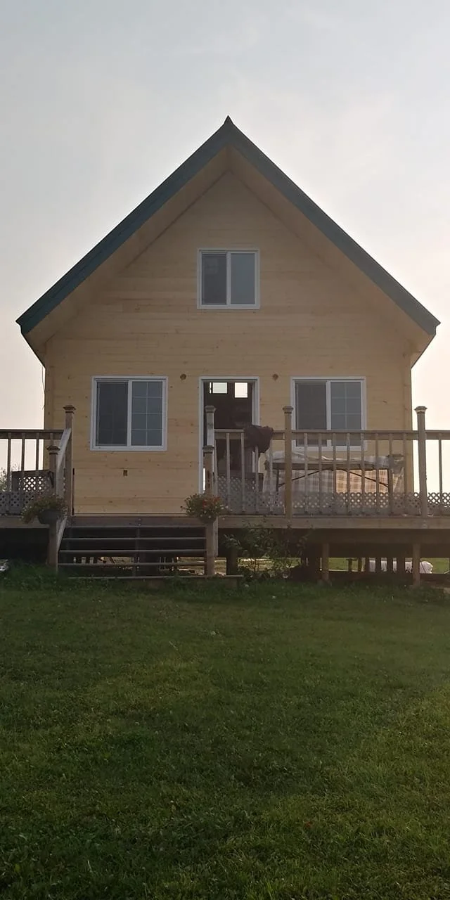 A two-story wooden house with a large front porch and a steep gabled roof. The house has three visible windows and a small staircase leading up to the porch.