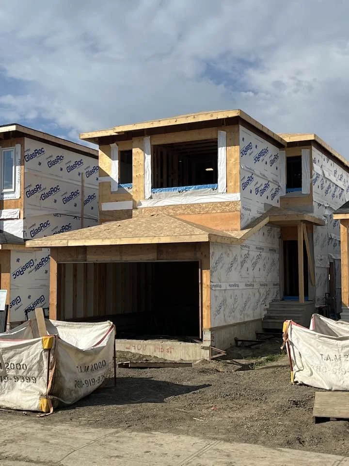 Under construction two-story house with plywood framing and weatherproof wrap, lacking exterior walls, with a front porch and stairs, on a partly cloudy day.