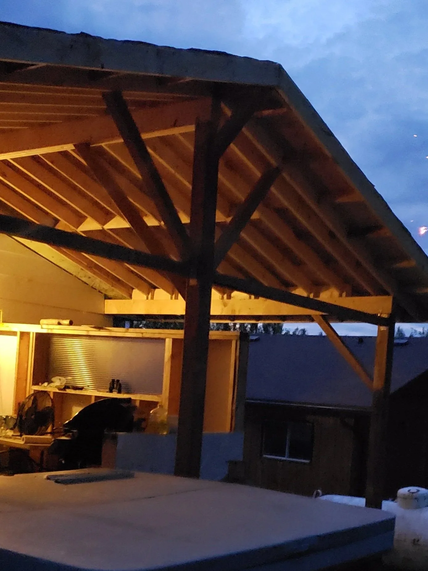 Construction site of a wooden structure, possibly a porch or deck, with visible beams, partially completed roof, and some construction tools and materials inside, during dusk.