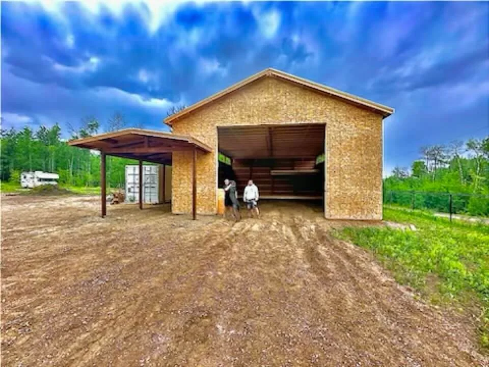 A large, partially constructed garage with a gable roof and open front, situated on a dirt lot with two people standing in front. There is a green landscape with trees and a cloudy sky in the background.