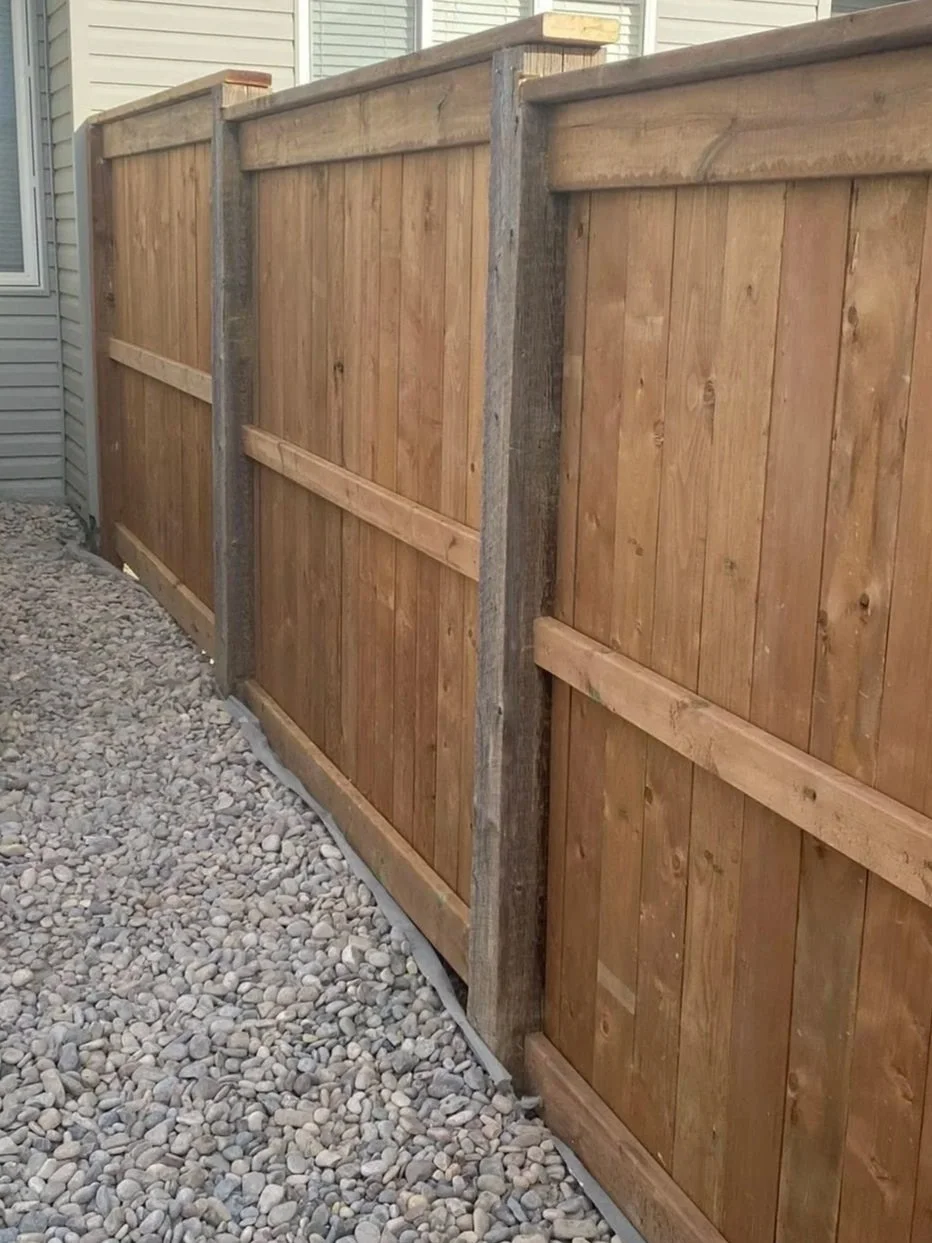 A newly constructed wooden privacy fence with vertical planks and horizontal support beams along a pebble-filled ground next to a house.