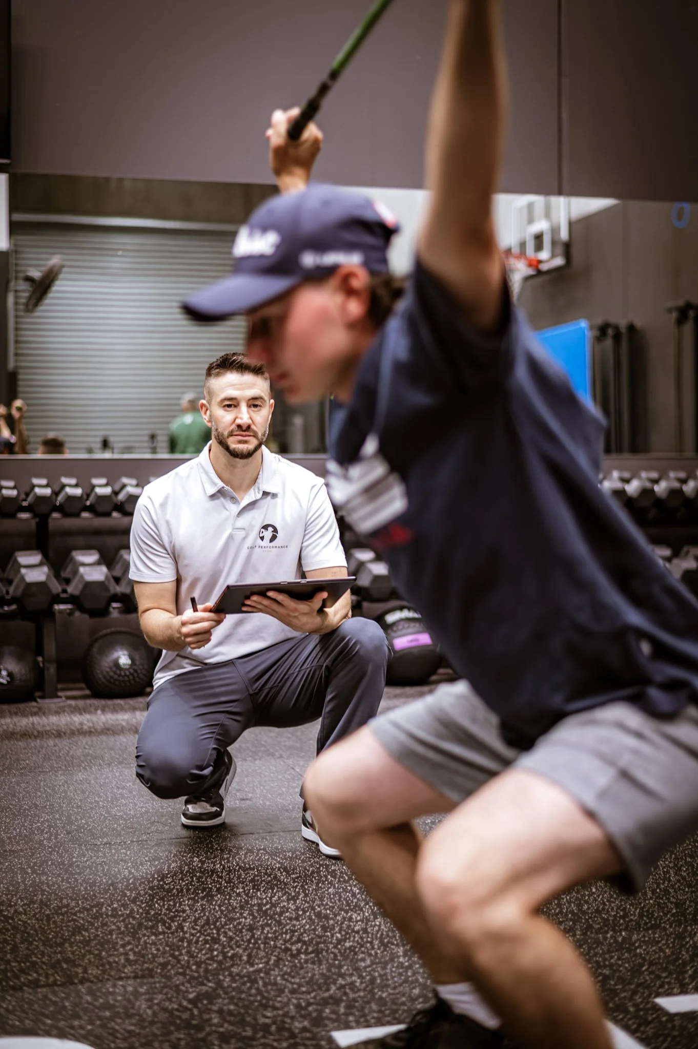 Personal trainer coaching a man doing squats while holding a racket in a gym.