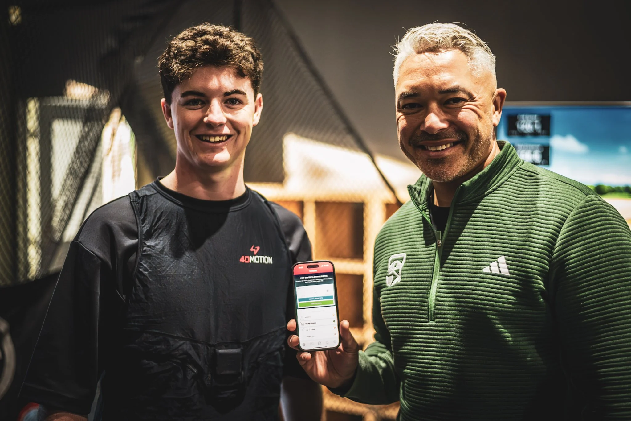 Two men smiling indoors, one holding a smartphone displaying an app, with gym or sports space in the background.