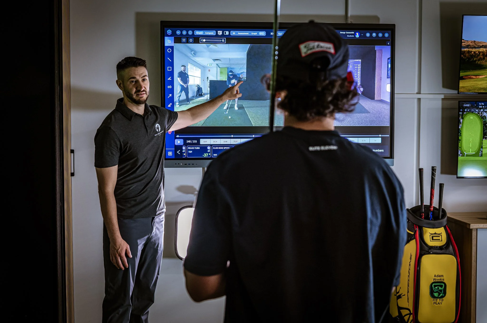 A golf instructor explaining a golf swing to a student in front of a large screen displaying golf swing analysis, with golf equipment nearby.