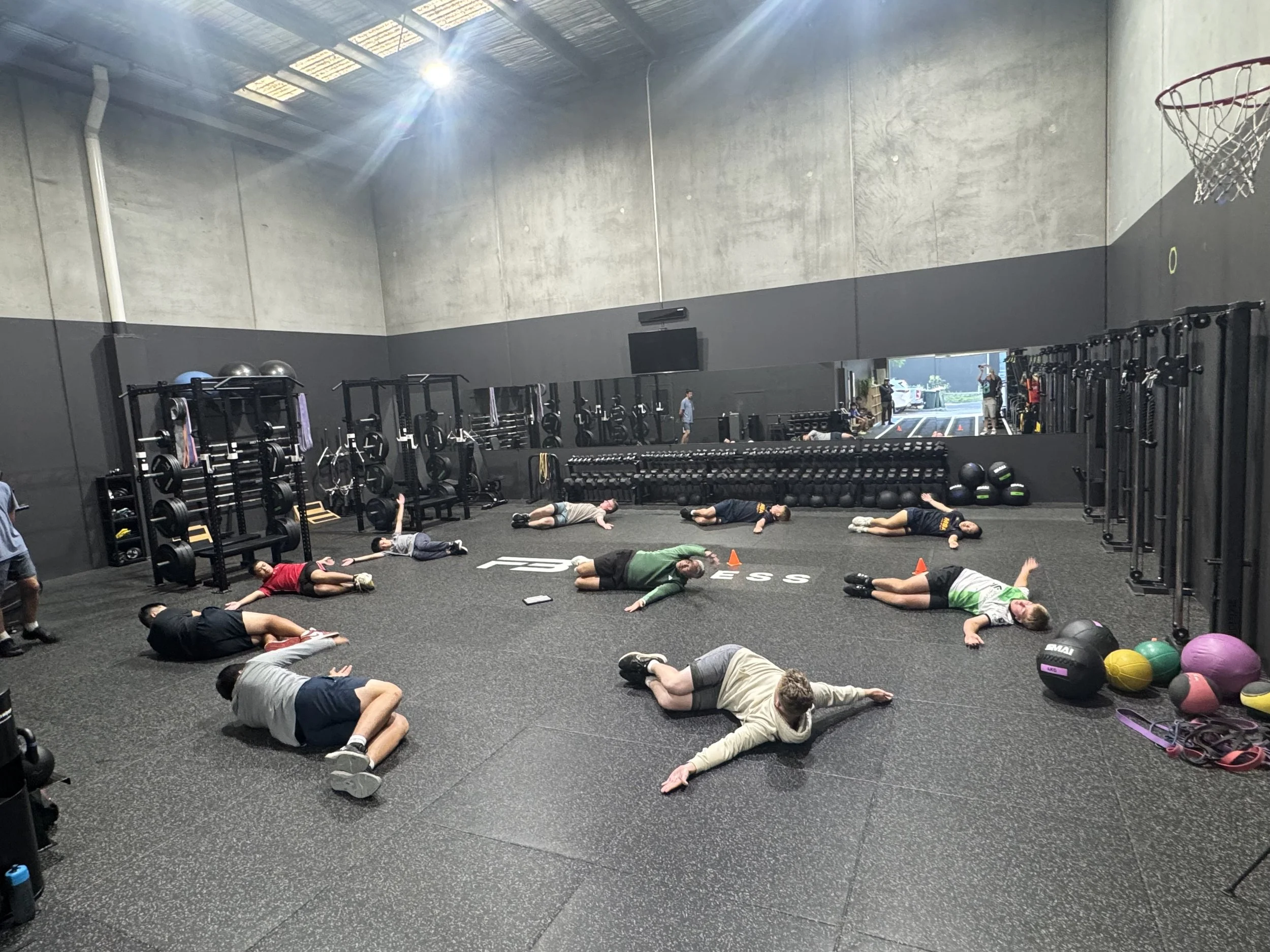 People lying on the gym floor during a fitness class or stretching session in a spacious gym with gym equipment and a large mirror.