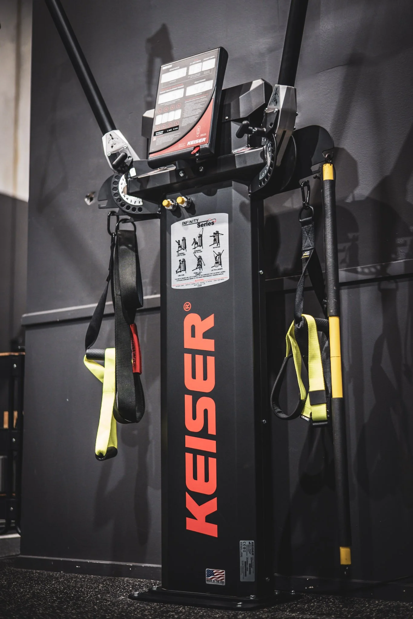 A black KEISER Infinity Series fitness machine with a large screen, attached resistance bands, and a yellow and black pole, set against a gray wall in a gym.