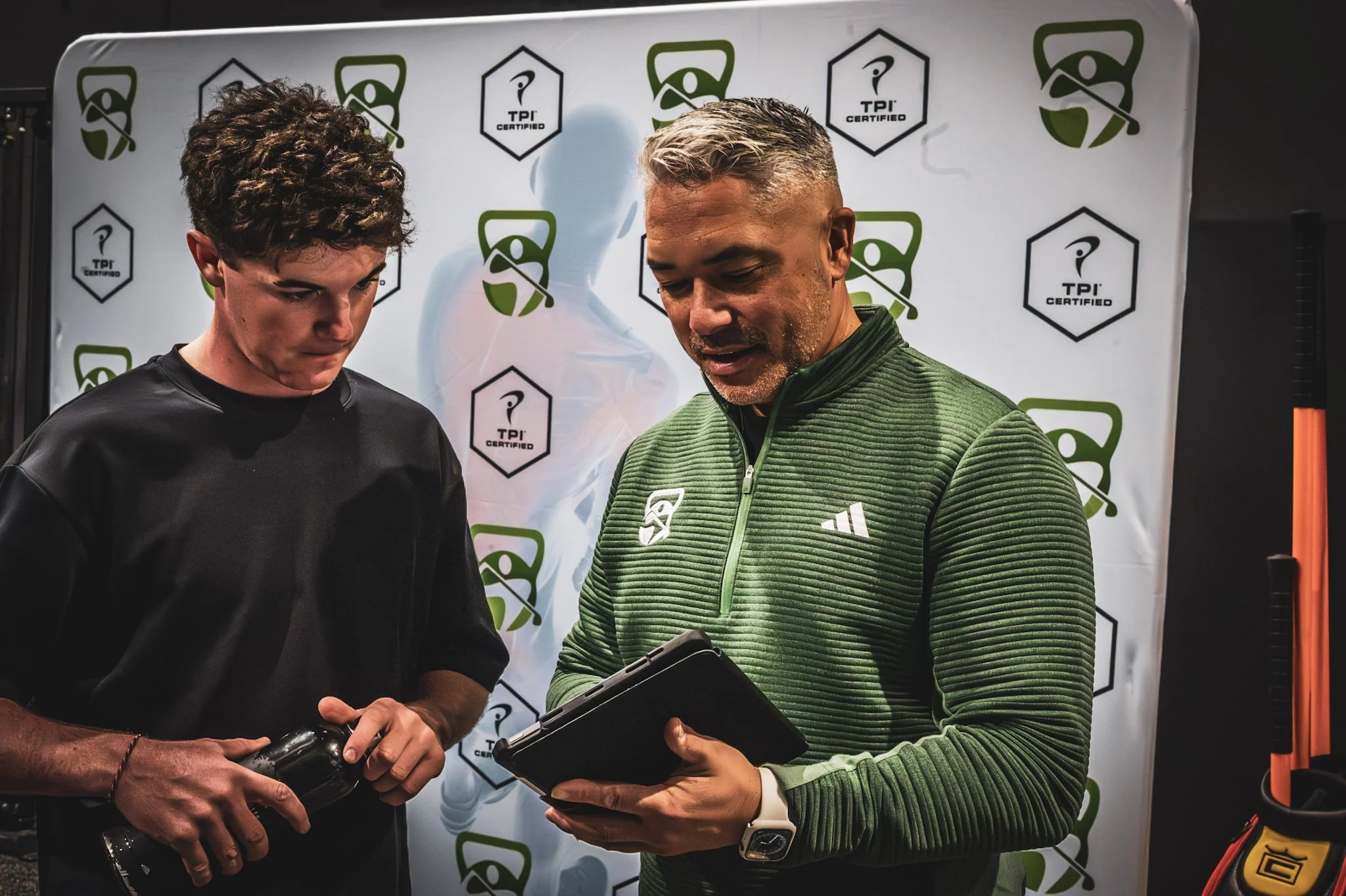 Two men, one younger and one older, standing in front of a backdrop with green and black logos that include a hexagon with a question mark and the words "TPI Certified." The younger man on the left has curly hair and is holding a water bottle. The older man on the right has short gray hair, is wearing a green zip-up jacket with brand logos, and is looking at a tablet he is holding.