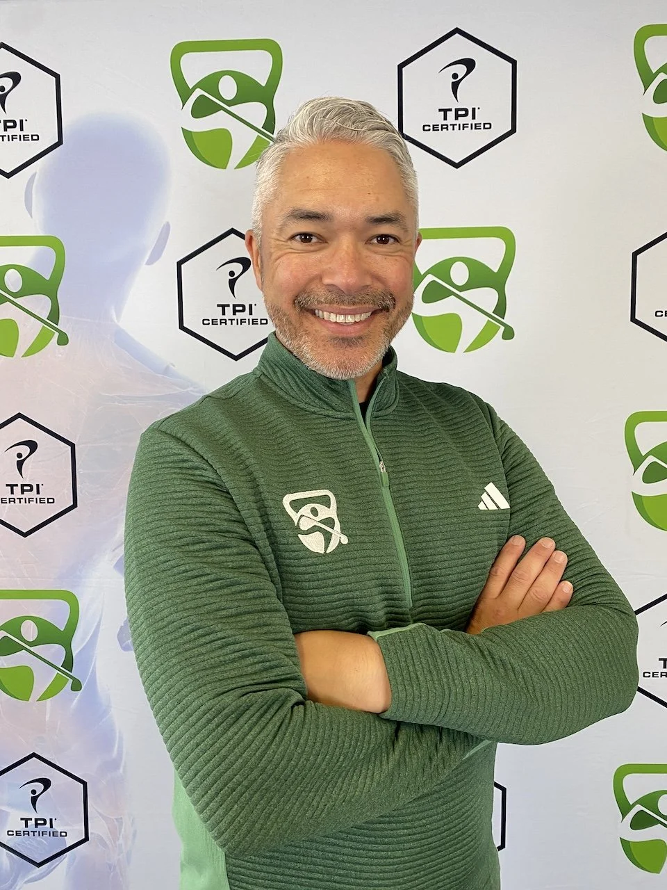 A smiling man with gray hair, short gray beard, wearing a green quarter-zip athletic jacket with logos, standing with arms crossed in front of a backdrop with green and black graphics and logos related to golf and TPI certification.
