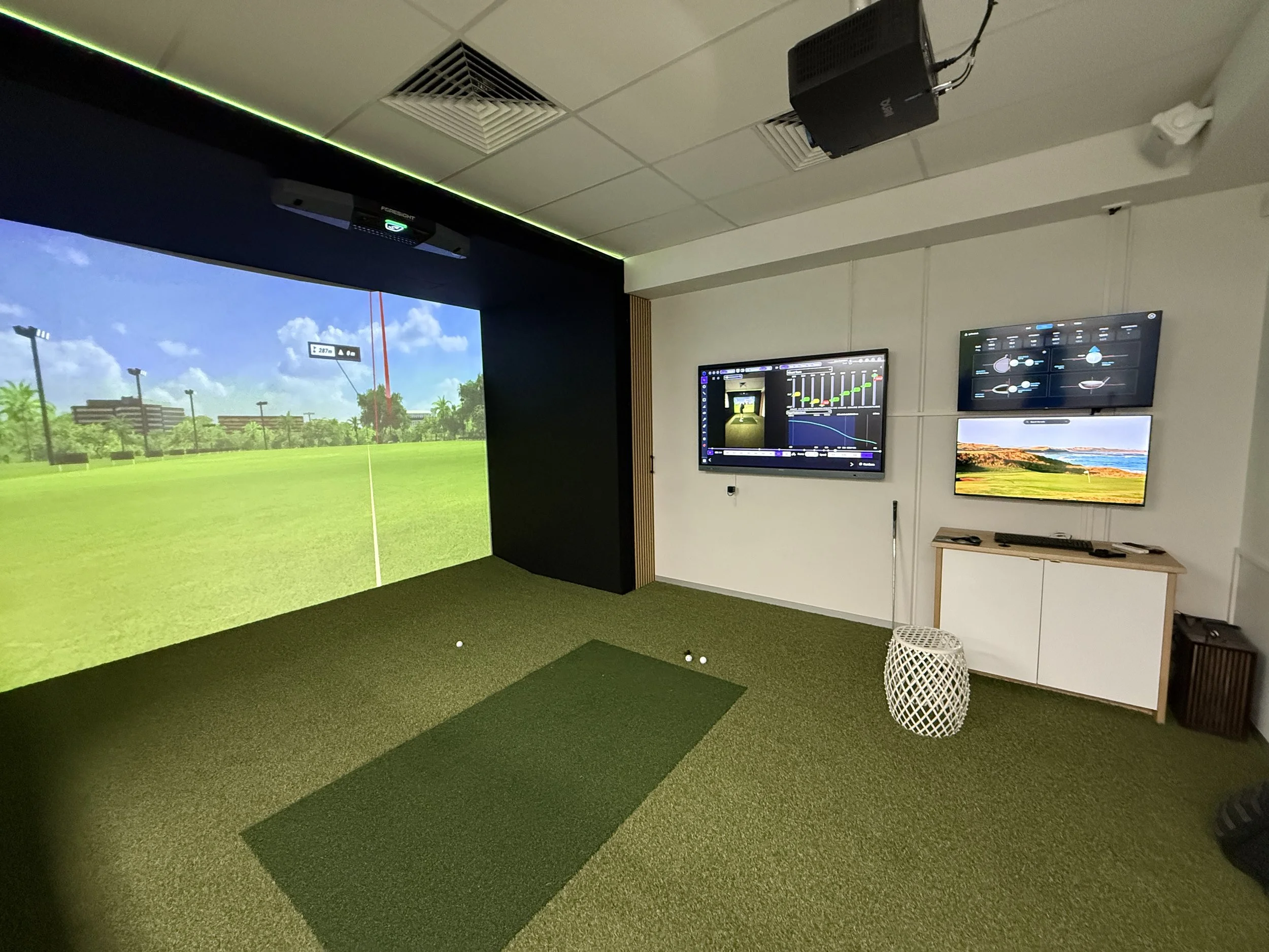 Indoor golf simulation room with a large screen displaying a golf course, a putting mat, and several monitors showing data and graphics.