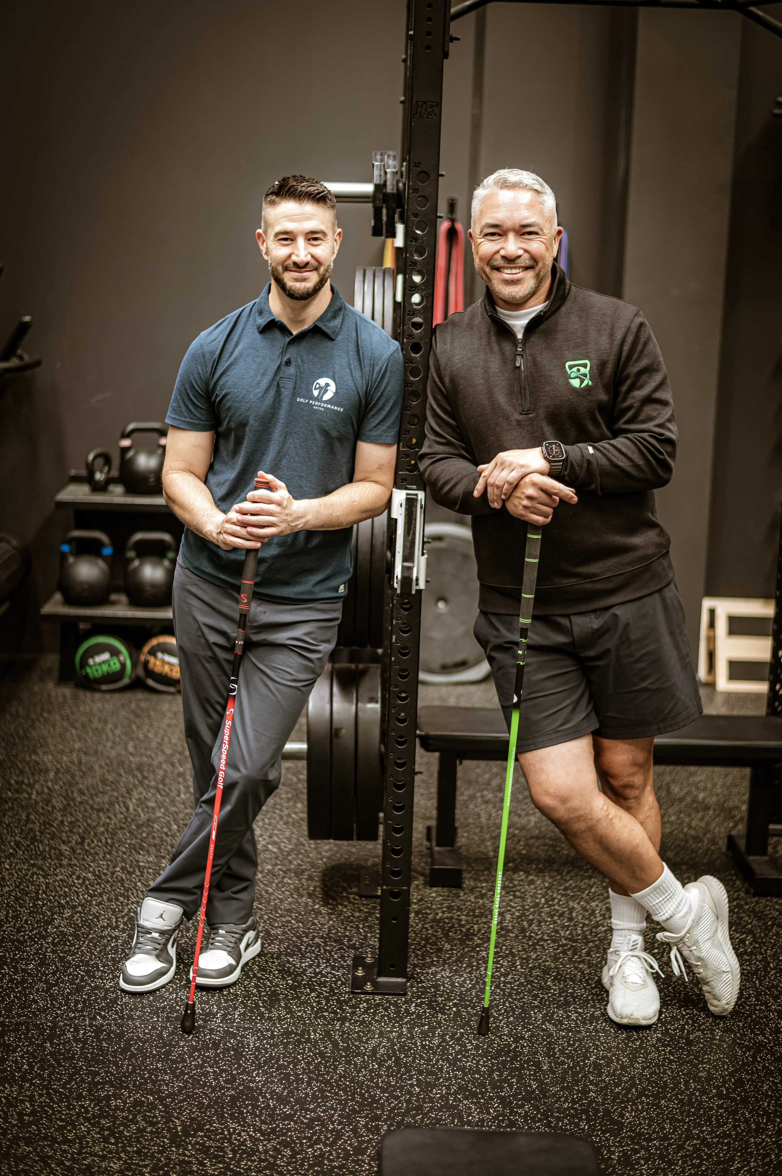Two men standing side by side in a gym, each holding a golf club, smiling at the camera. The gym has black walls and equipment like kettlebells and weight plates in the background.