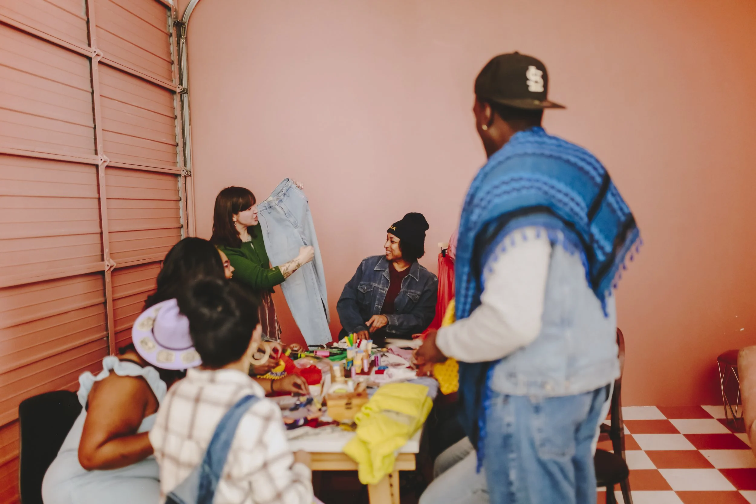 Group of people at a craft table with clothing and art supplies, indoors with pink walls.