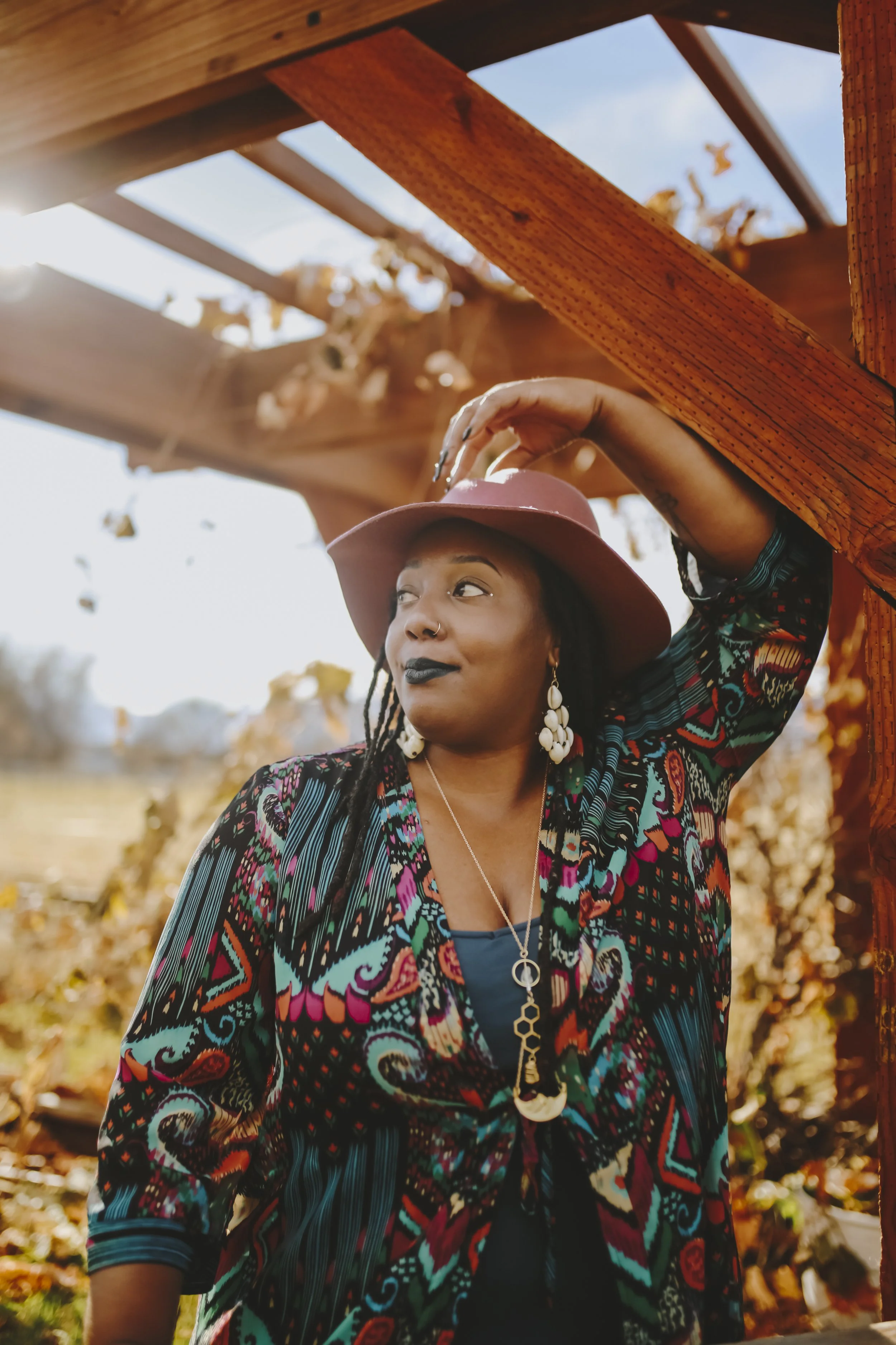 Nyasha Williams in dark lipstick, wearing a large pink hat, colorful patterned jacket, and jewelry, stands outdoors under a wooden structure with falling leaves and clear sky in the background.