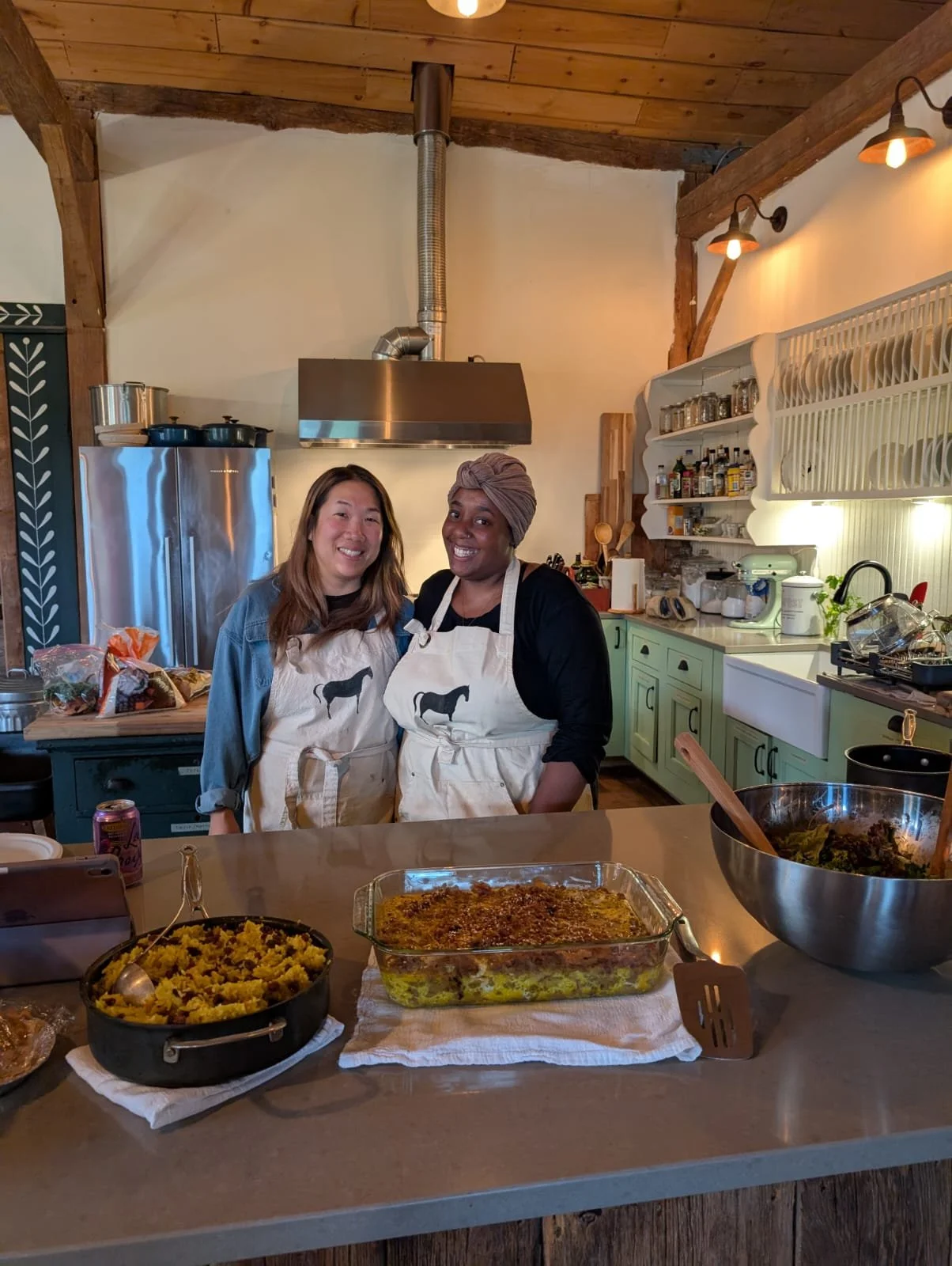 Two women standing in a kitchen, smiling, wearing aprons with a horse design. The kitchen has green cabinets, open shelves with dishes, and a stainless steel refrigerator. There are two prepared dishes on the counter in front of them.