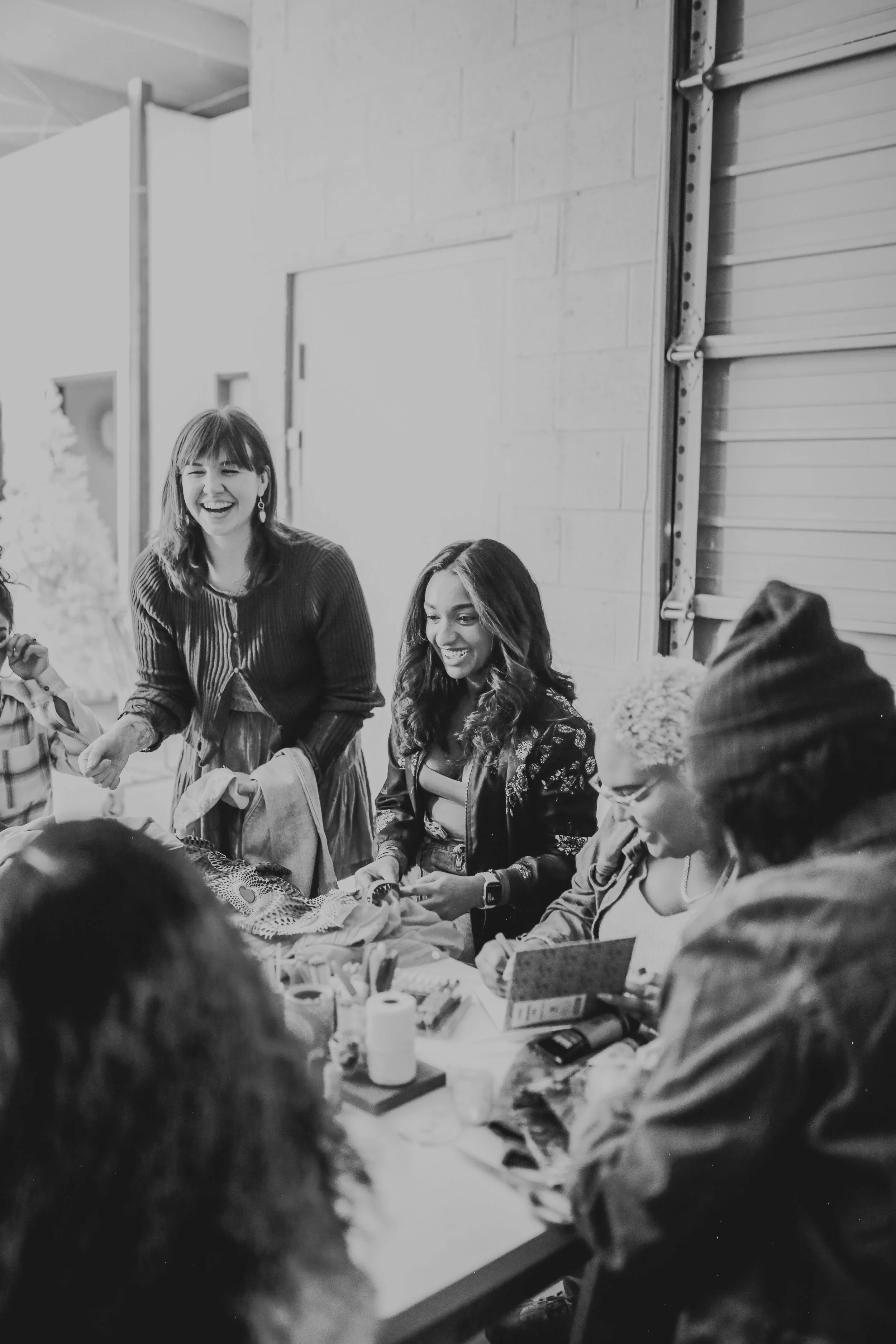 A group of women gathered around a table, smiling and engaging in conversation, in an indoor setting.