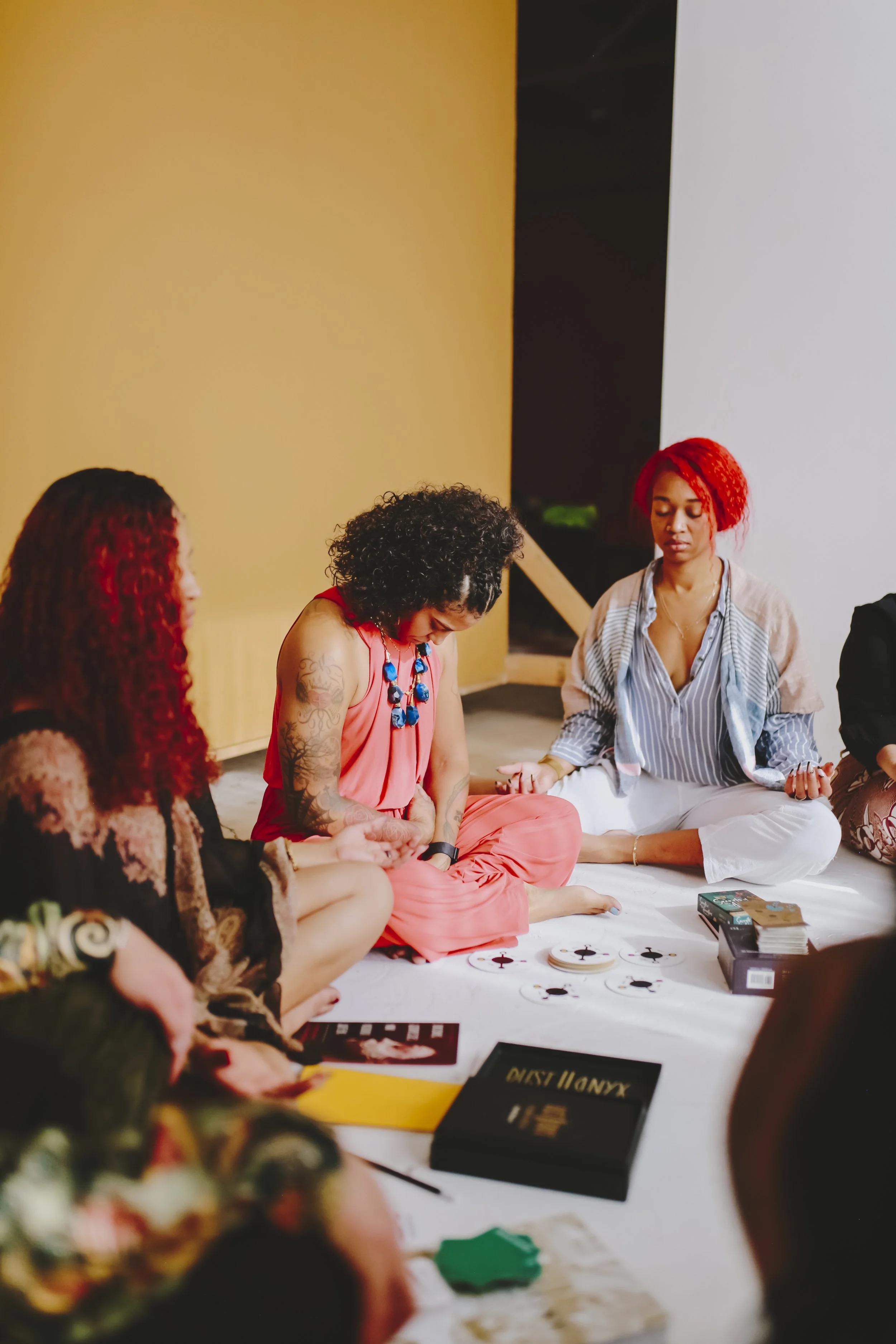A group of women sitting on the floor engaged in a tarot card reading or discussion, with tarot cards and related items on the white cloth in front of them.