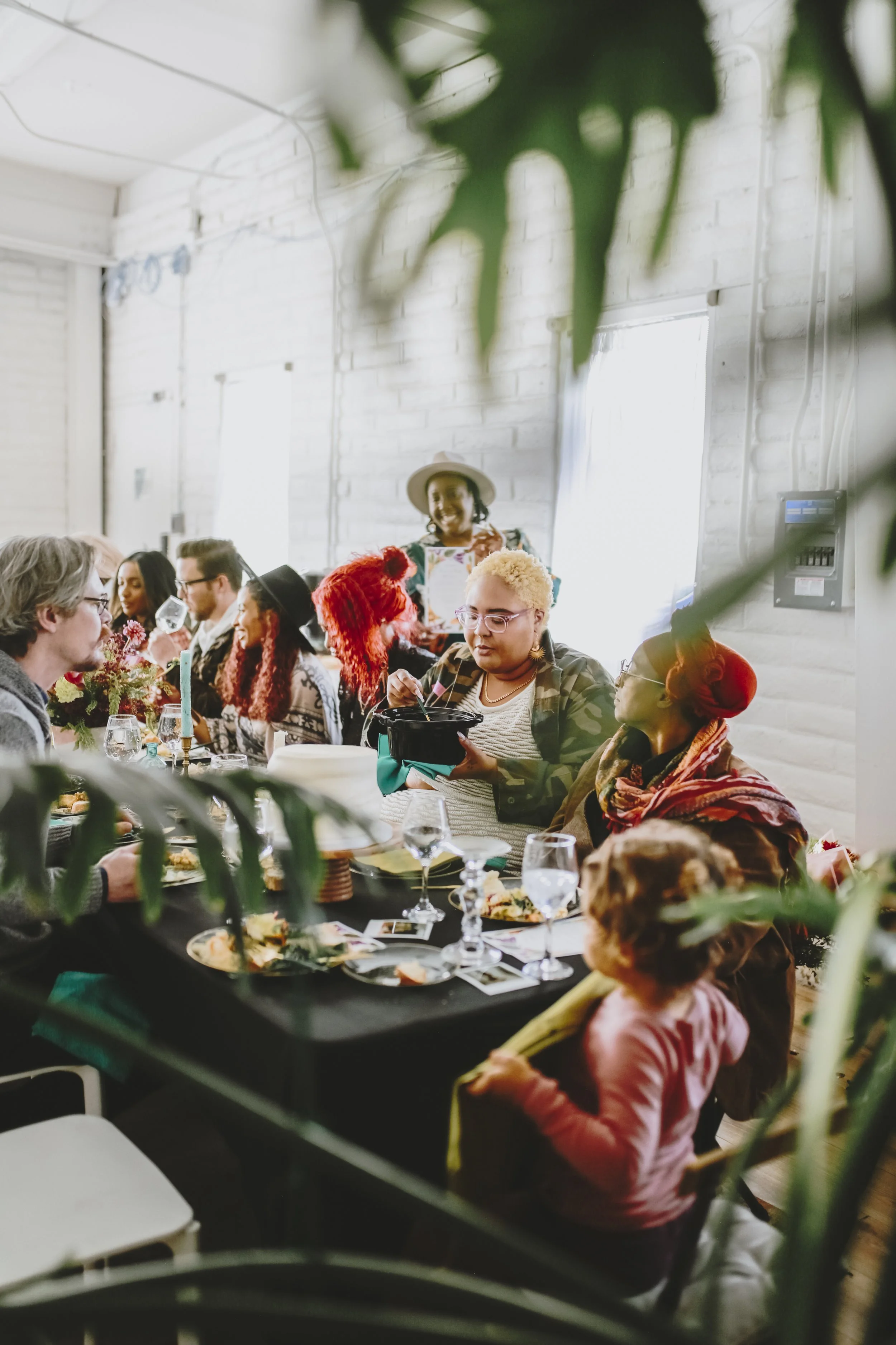 People gathered at a dinner party, seen through green leaves, with a group of diverse individuals dining together indoors, some talking and some eating, in a bright room with white brick walls.
