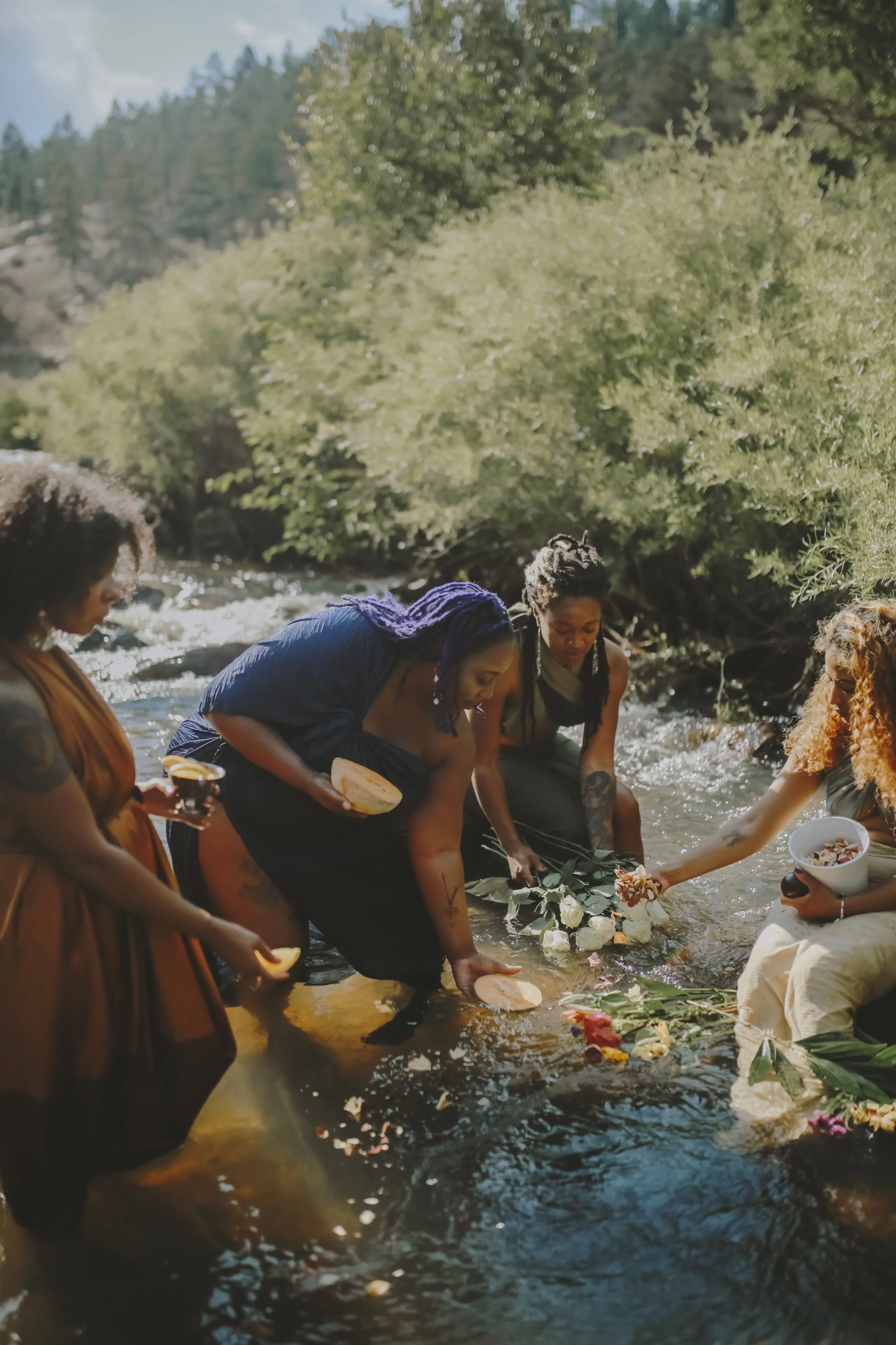 A group of women with diverse hairstyles and tattoos gather in a river, holding flowers and bowls, as part of a ritual or ceremony against a lush green landscape with trees and hills in the background.