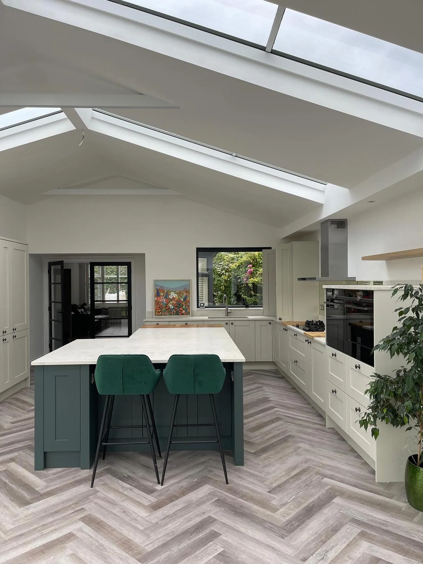 Modern kitchen with skylights, white cabinets, a kitchen island with green chairs, a window with greenery outside, and light wood herringbone flooring.