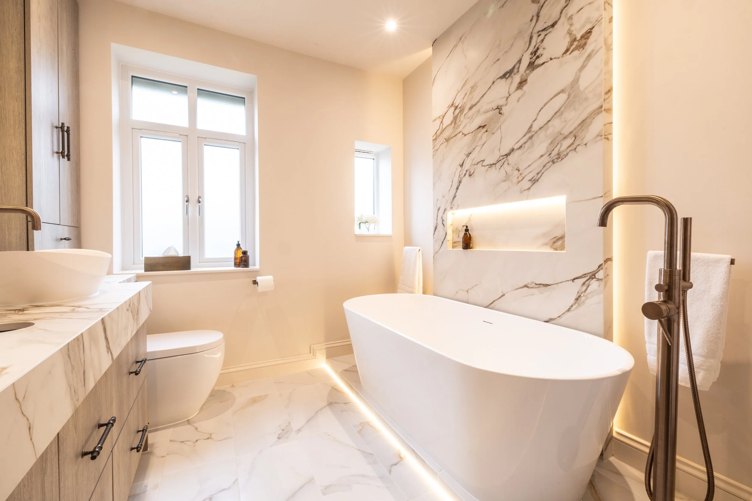 Modern bathroom with a large white bathtub, marble walls and floor, wooden cabinetry, a vessel sink, a window with natural light, and minimalist decor.