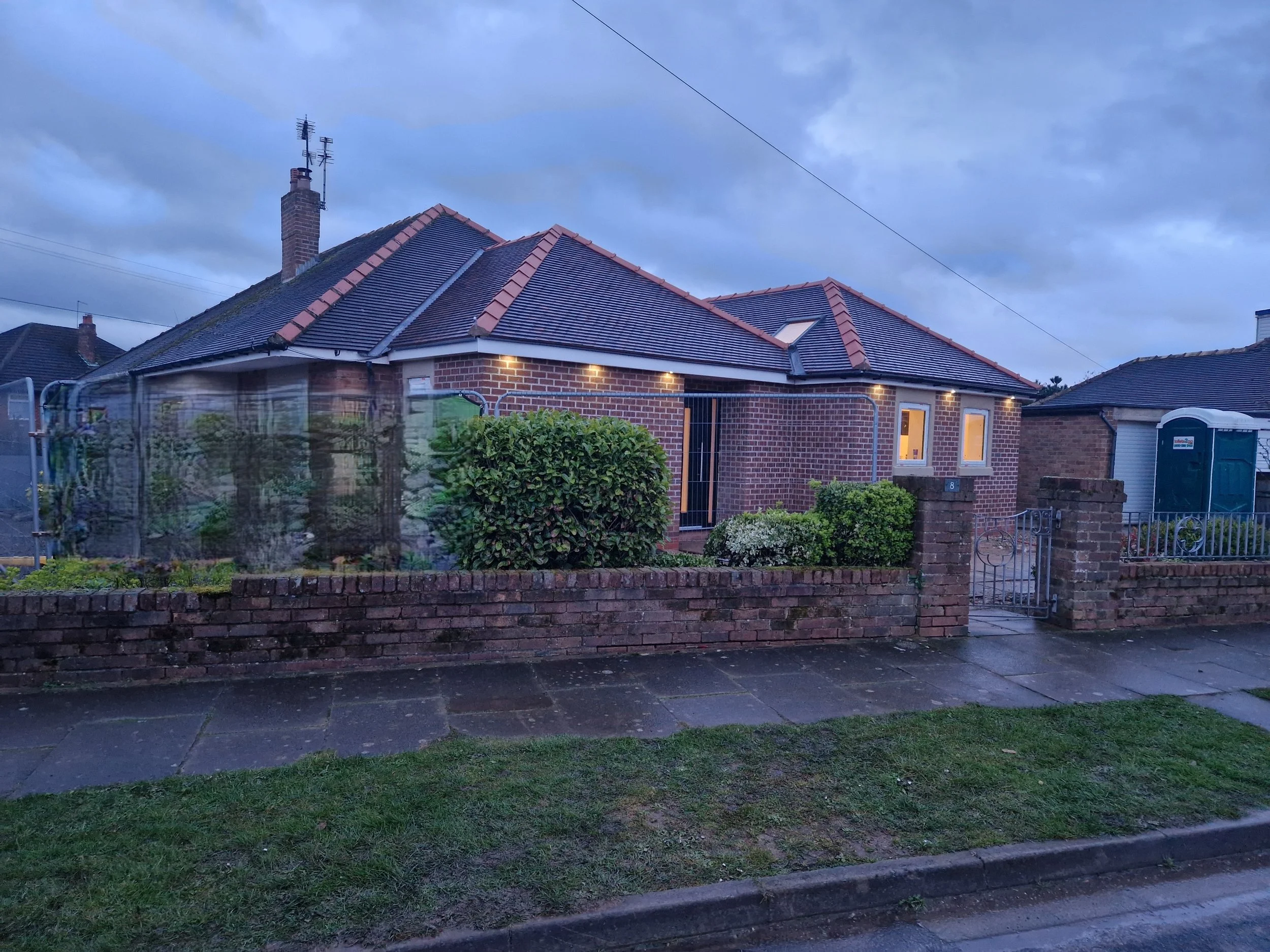 A house with a brick exterior, gabled roof, and a white-lit porch, enclosed by a brick fence with a small gate, and a garden with bushes. Overcast sky in the background.