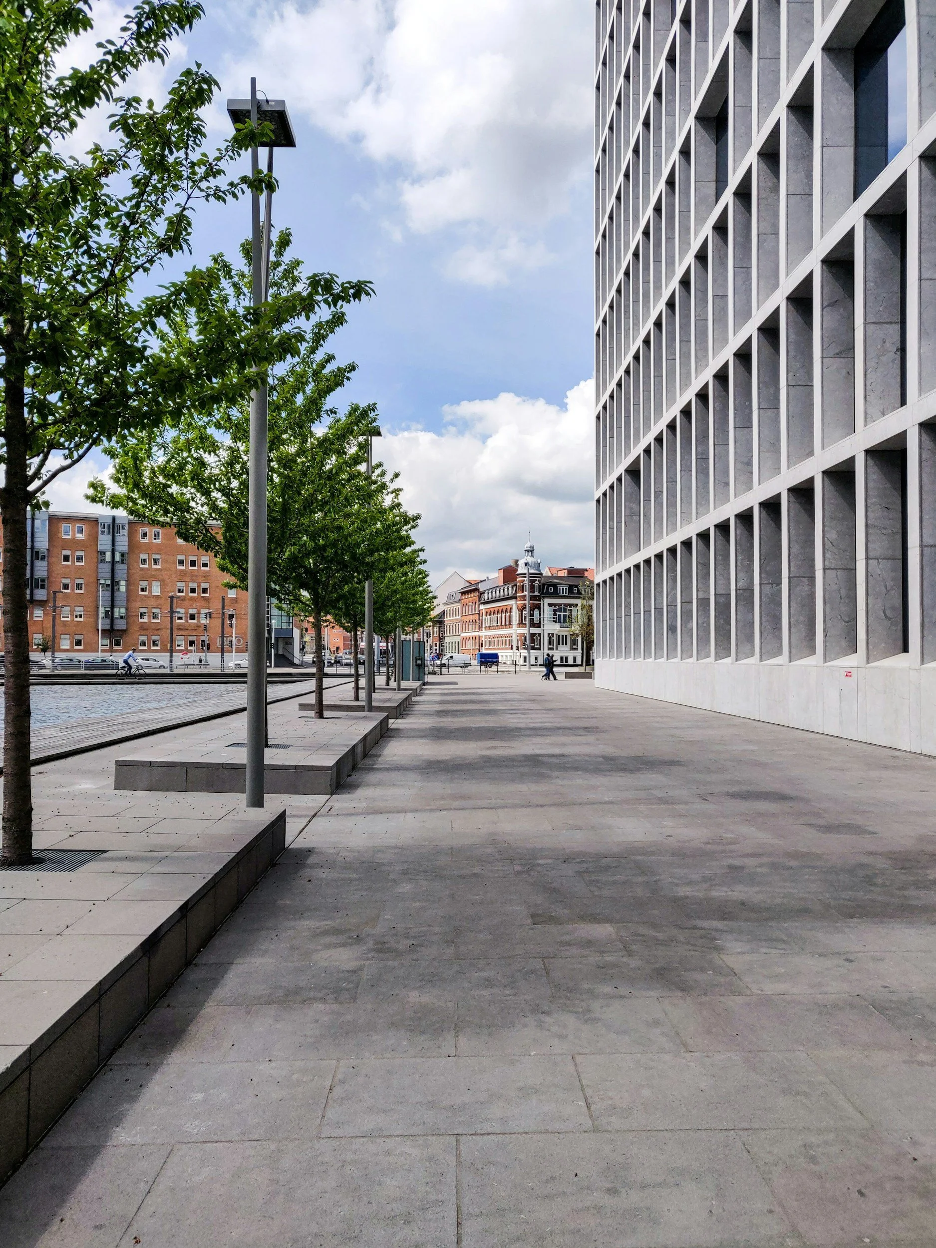 Empty city sidewalk with new trees, modern street lamps, and a large glass-fronted office building on the right under a partly cloudy sky.