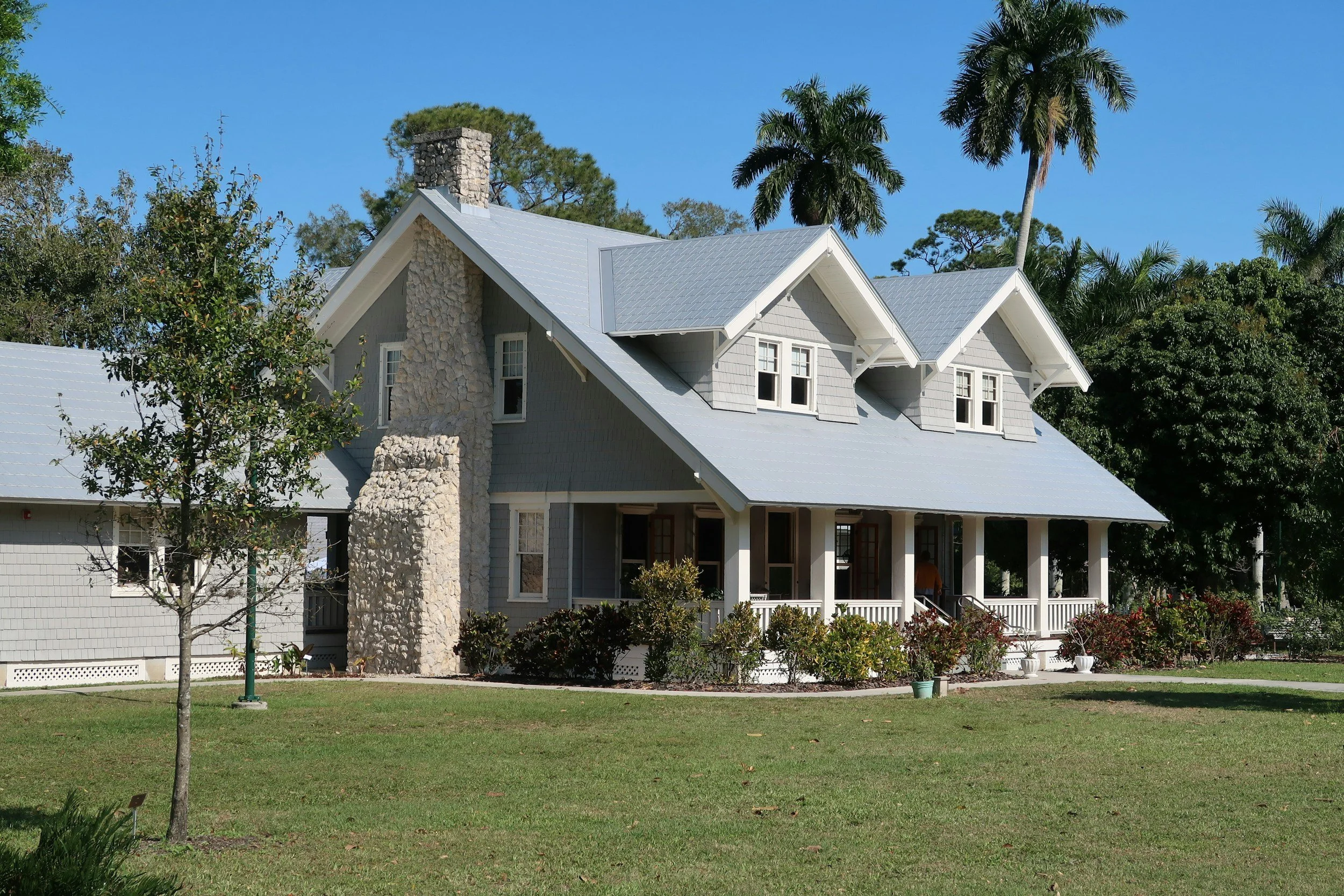A large gray two-story house with a steep metal roof, multiple windows, a front porch with white railings, and a stone chimney, surrounded by trees and a well-maintained lawn.