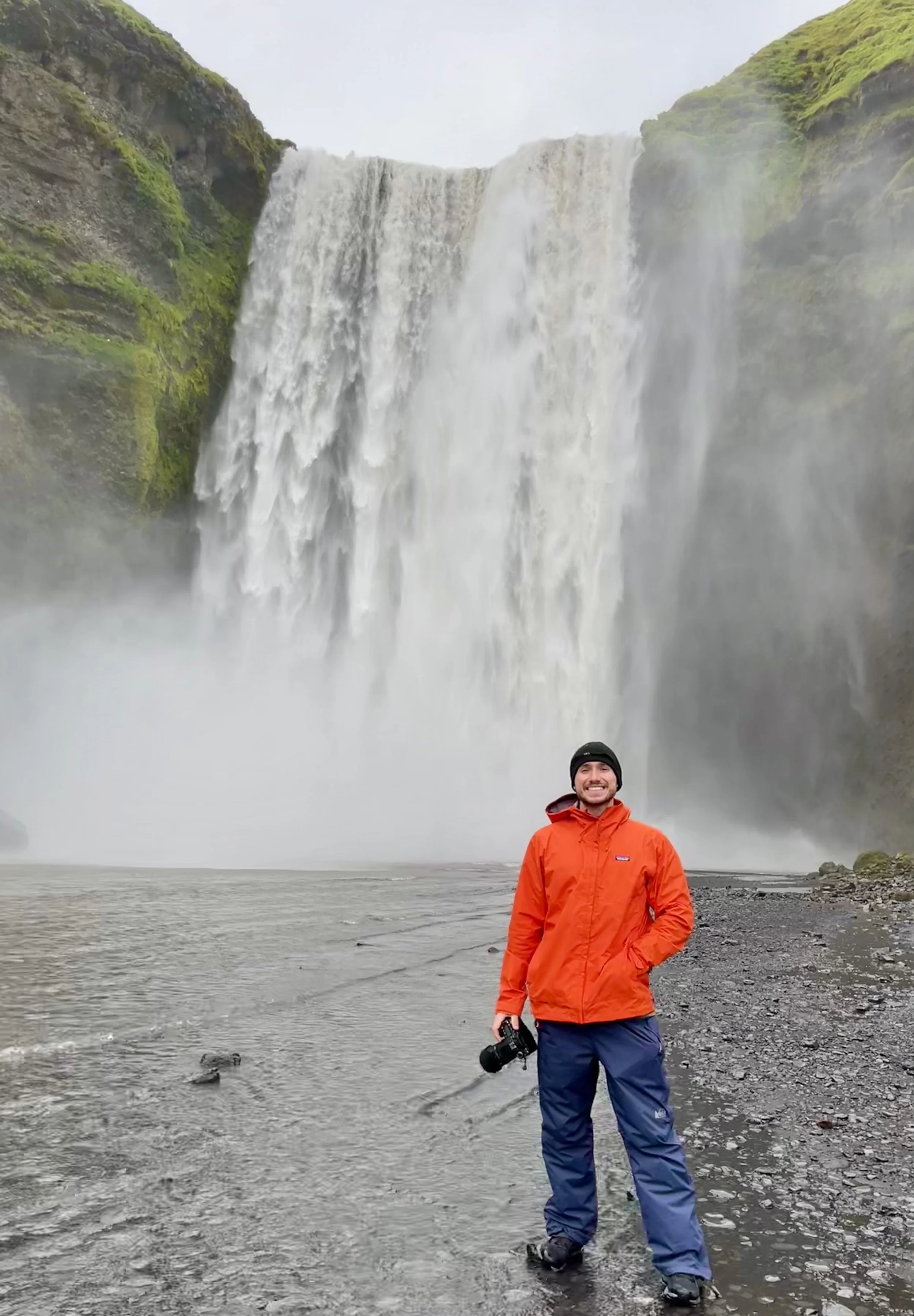 A person in an orange jacket and blue pants standing on a rocky, wet shore with a camera in hand, smiling in front of a large waterfall surrounded by green cliffs and mist.