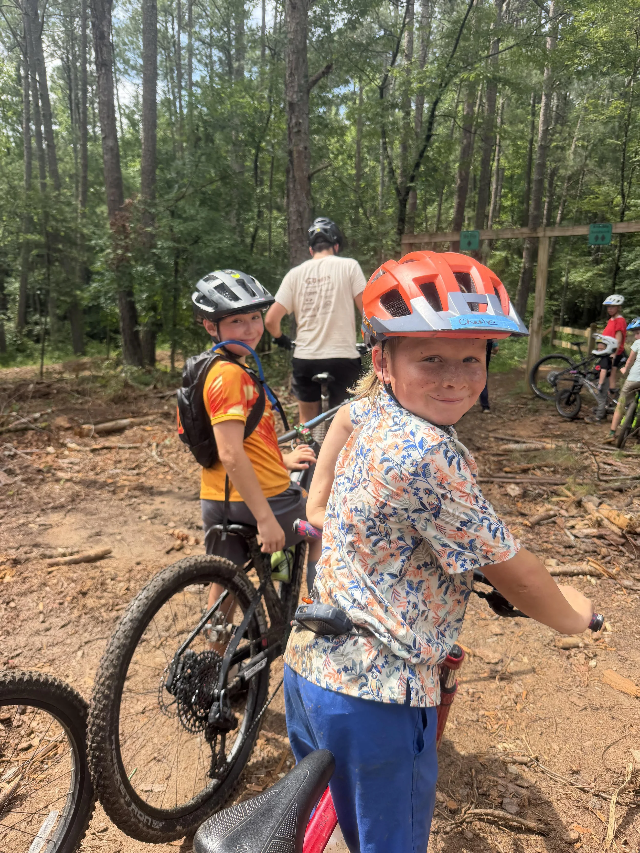 Children riding mountain bikes on a dirt trail in a wooded area with many trees, some children wearing helmets and smiling.