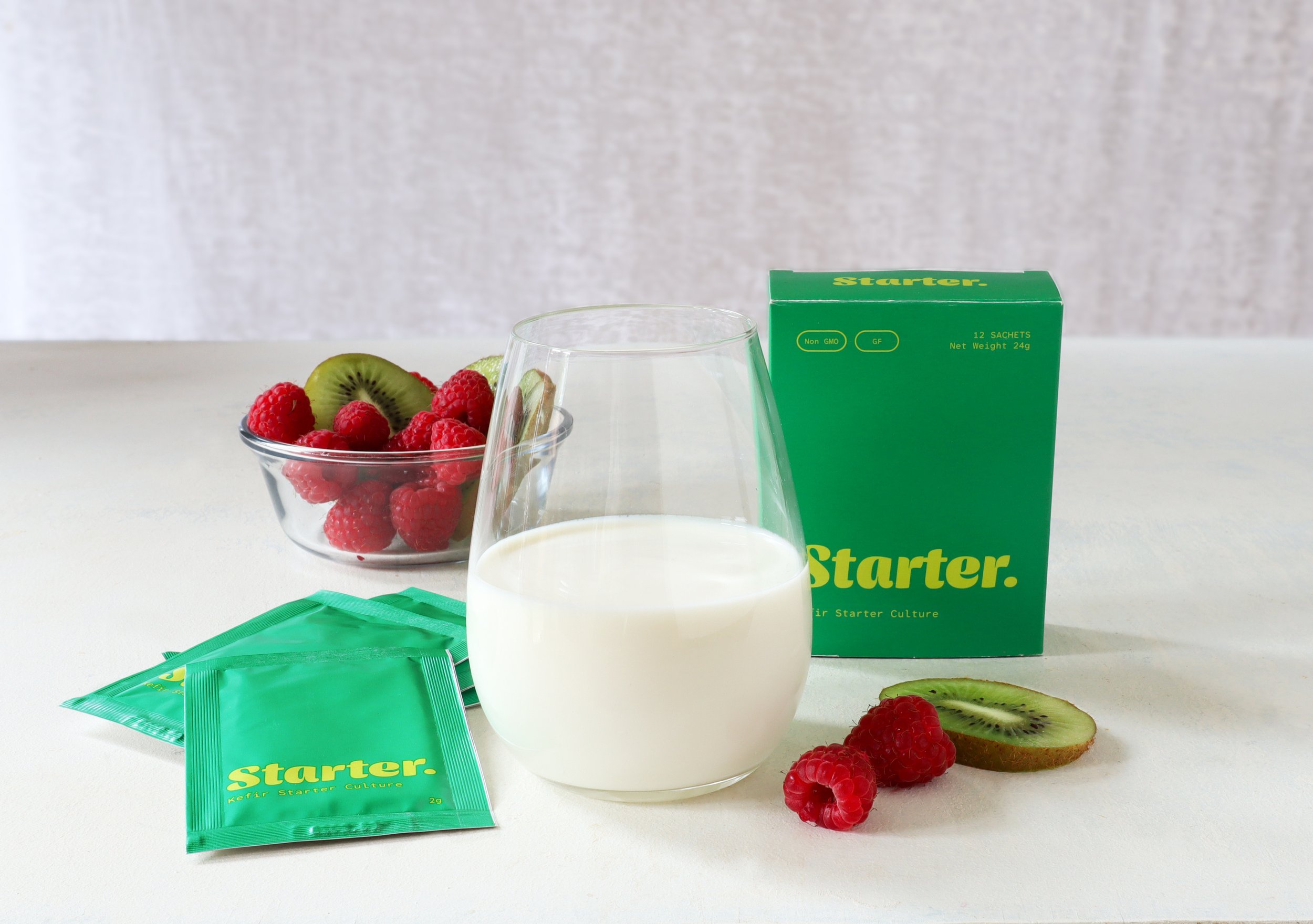 A glass of kefir or yogurt, a bowl of fresh raspberries and kiwi slices, and green starter culture packets on a white surface with a plain gray background.