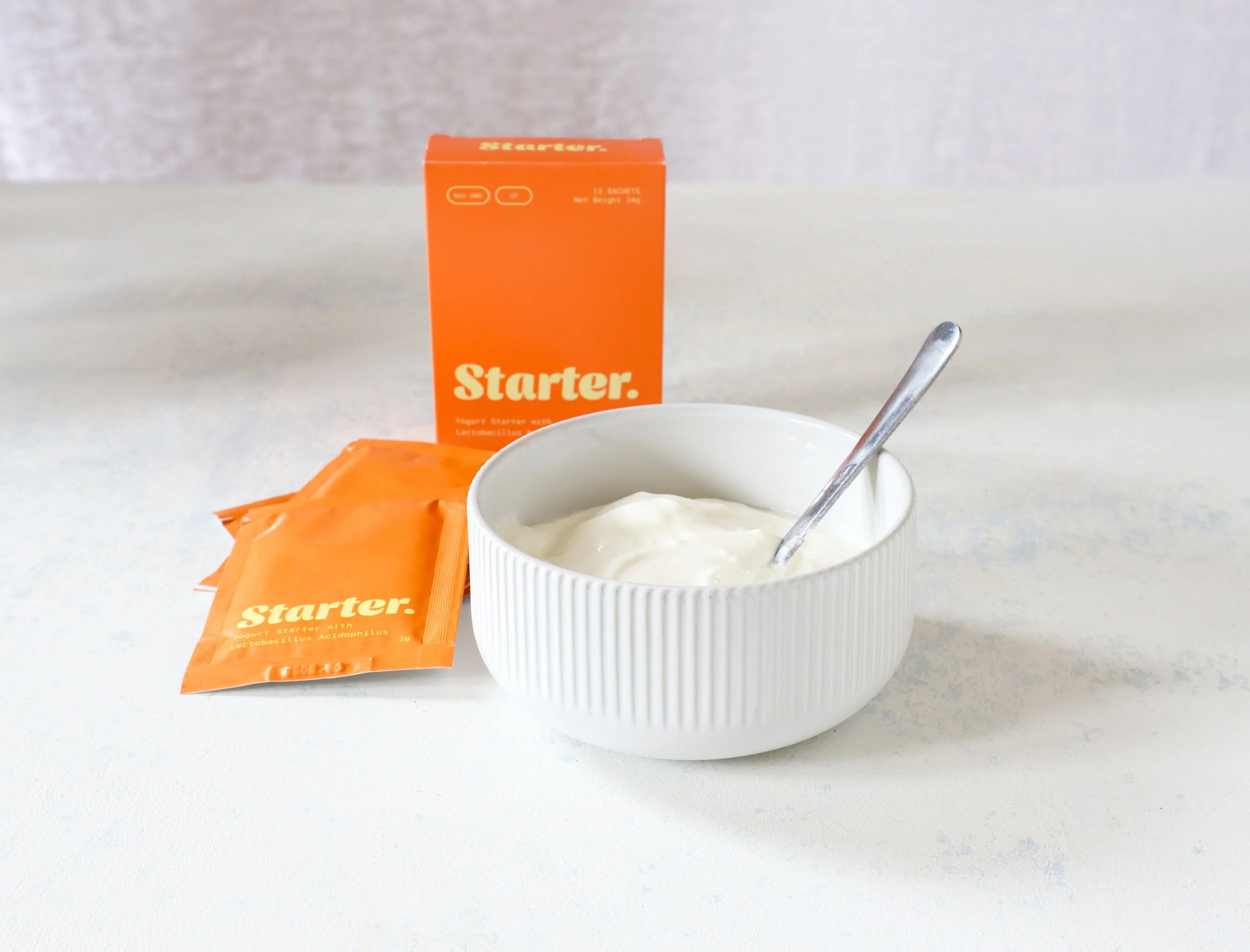 A small orange box labeled 'Starter' with text indicating it contains 12 sachets, set weight of 24g, and features probiotics. A white ceramic bowl filled with yogurt and a metal spoon, along with orange sachets, are set on a light-colored surface.